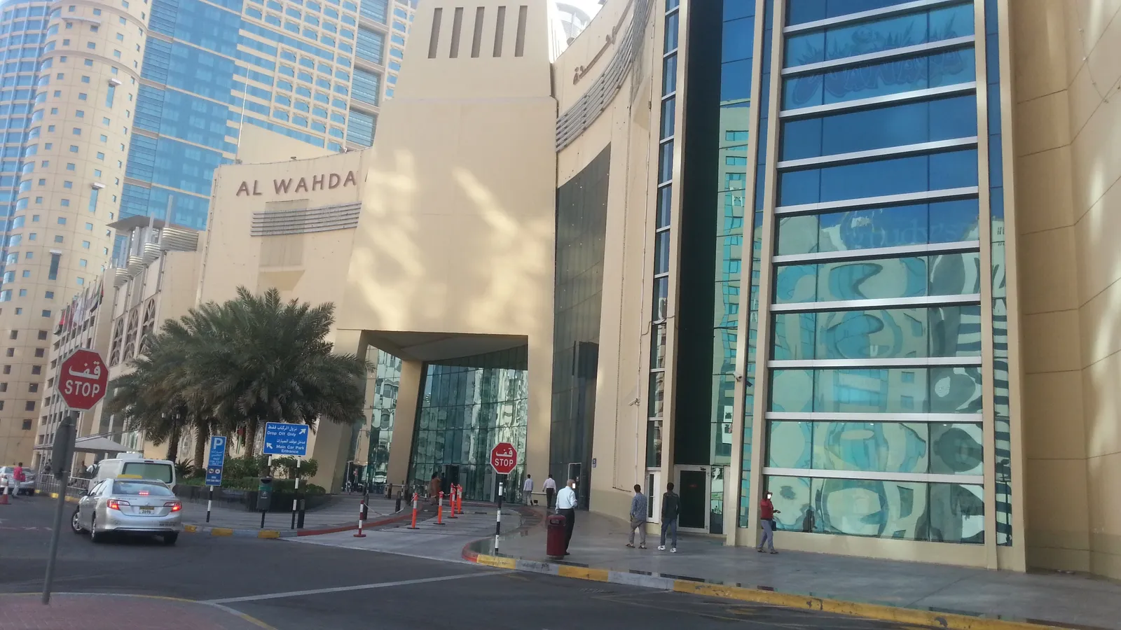 The glass and stone entrance of Al Wahda Mall in Abu Dhabi with its name on the facade, palms in the forecourt and office towers along the street beside it