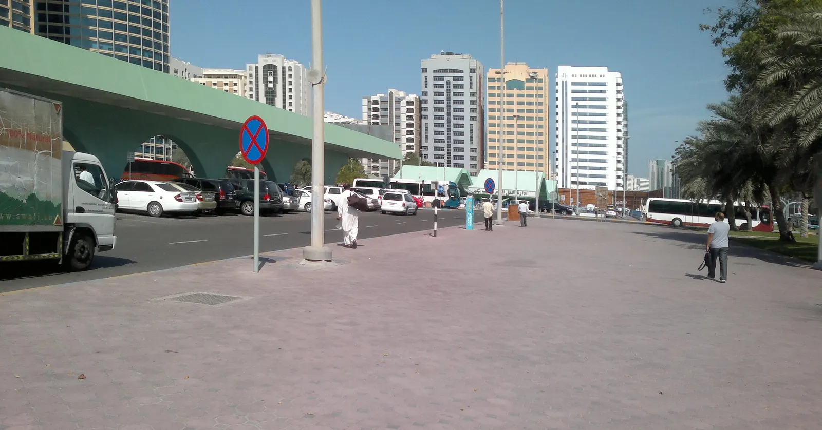 Coaches drawn up under a green canopy at Abu Dhabi's Central Bus Station, with passengers crossing the forecourt and residential towers rising behind
