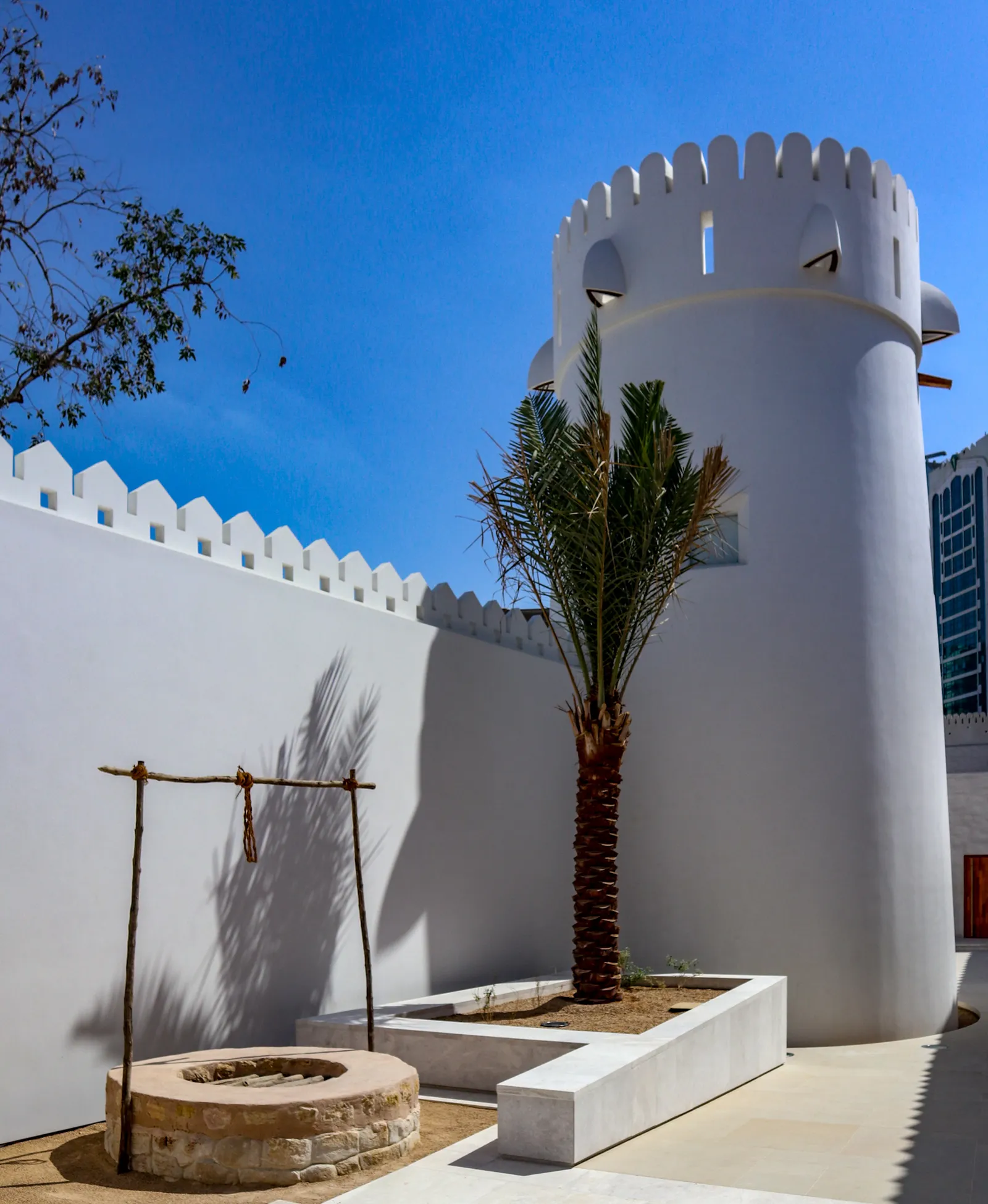 A stone well head with a wooden rope frame over it in the courtyard of Qasr Al Hosn, with the whitewashed crenellated wall and round tower of the fort behind and a date palm alongside
