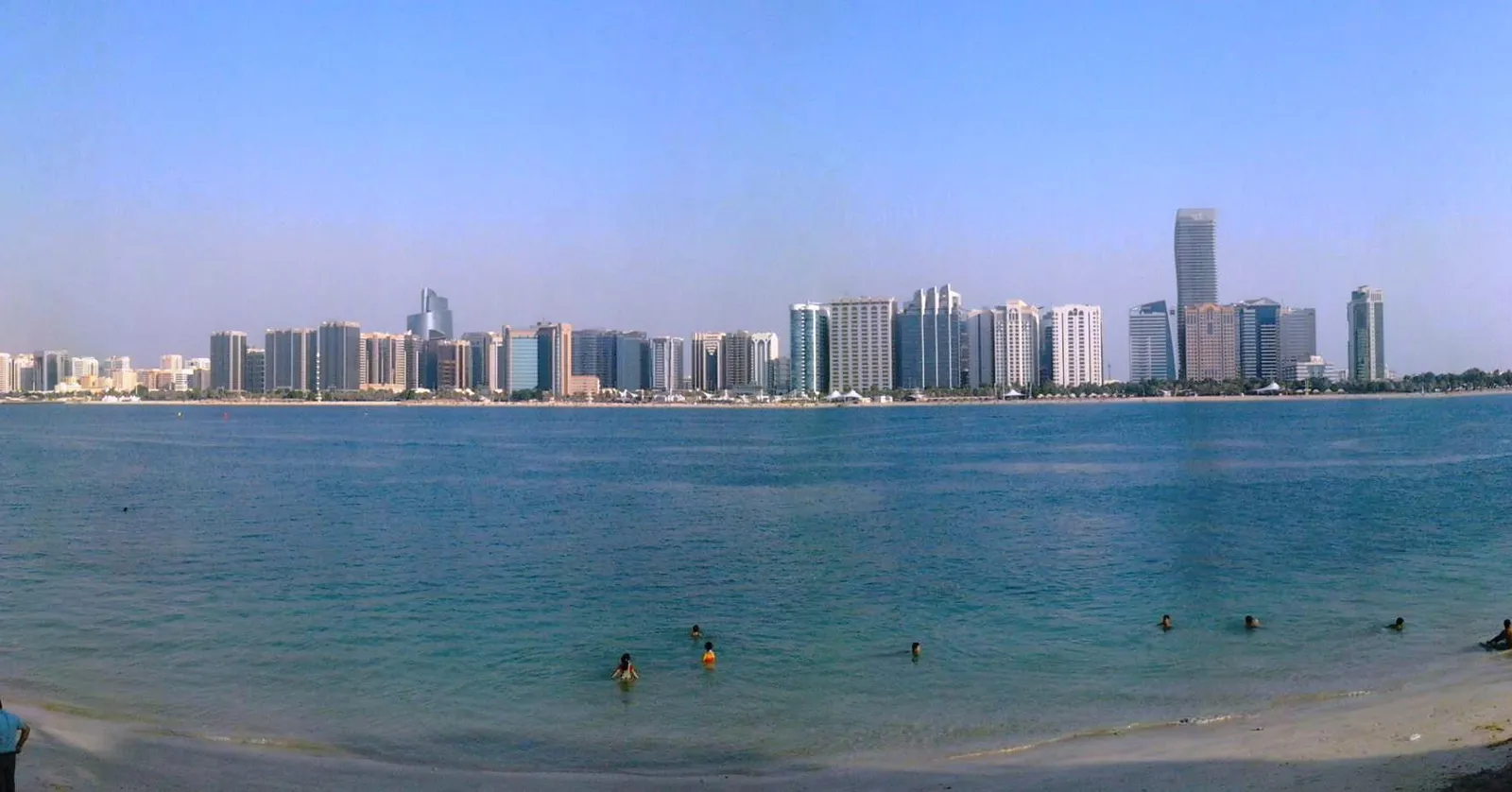 Swimmers in the shallow turquoise water off Corniche Beach in Abu Dhabi, with the line of white residential towers along the seafront behind them