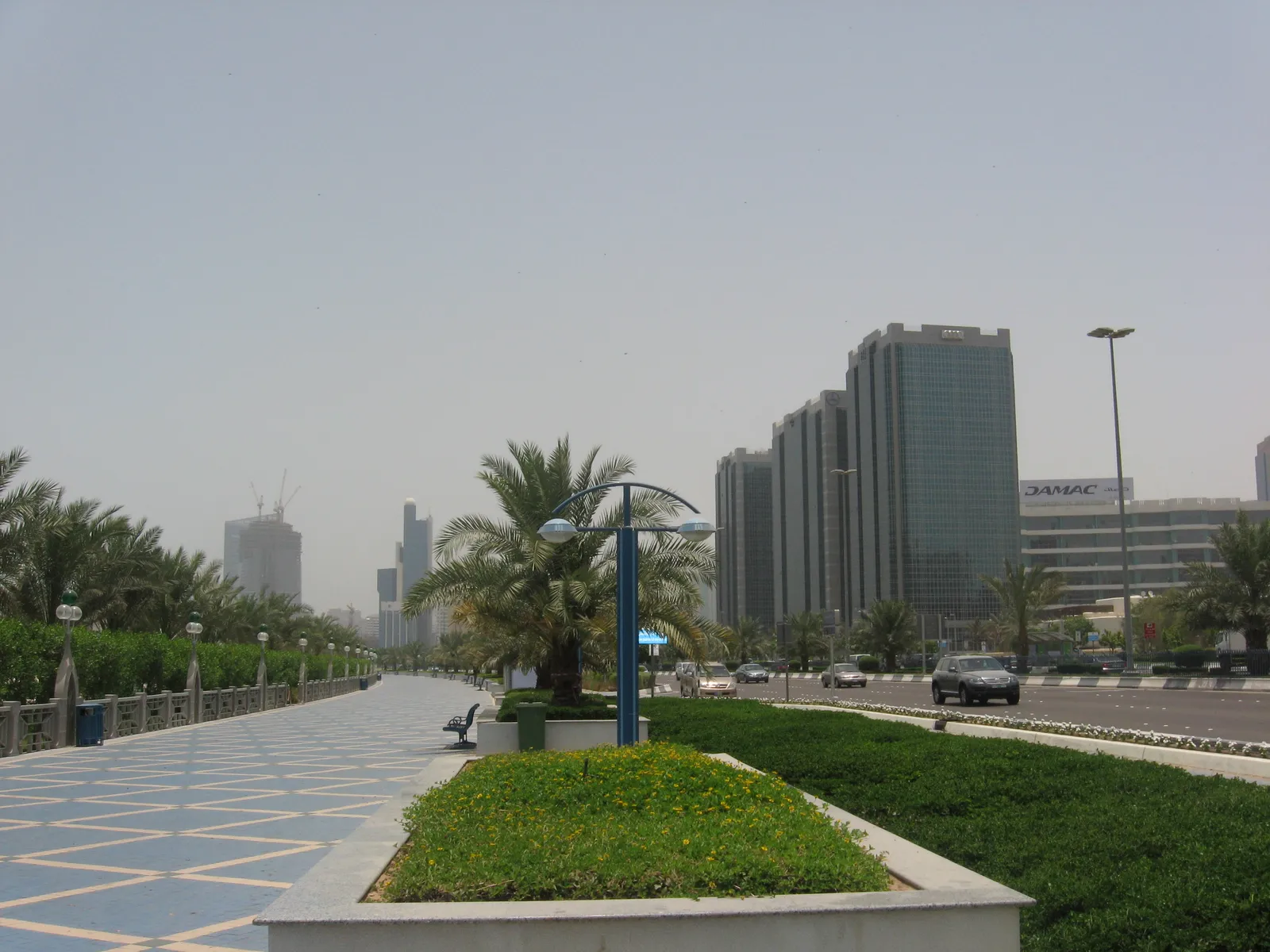 The blue-patterned paved promenade along Abu Dhabi's Corniche Road, with a planted median, young palms and lamp posts, and office towers on the inland side