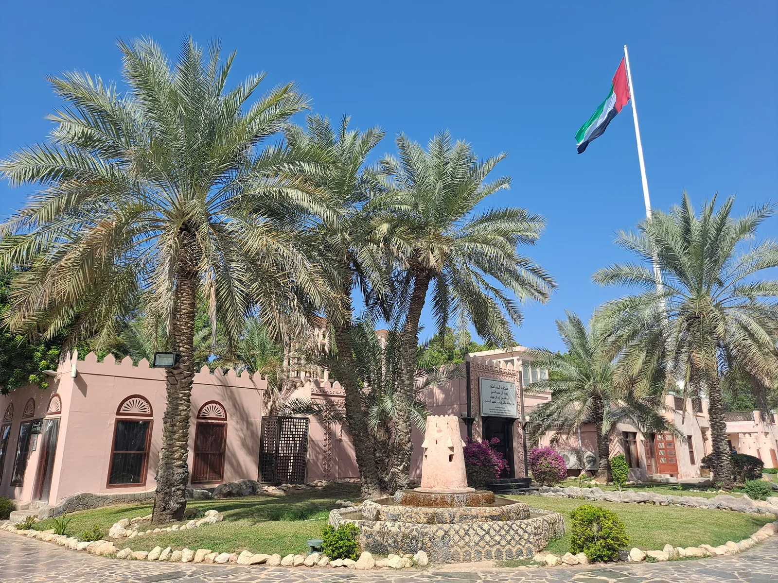 Low pink mud-brick buildings with crenellated walls and date palms at Abu Dhabi's Heritage Village, with the UAE flag flying from a tall pole behind them