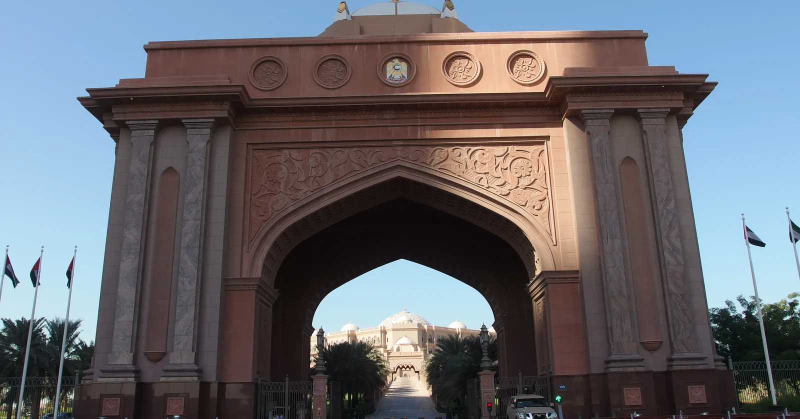 The monumental pink stone gateway arch at Emirates Palace in Abu Dhabi, with flagpoles either side and the domed palace hotel visible through the opening