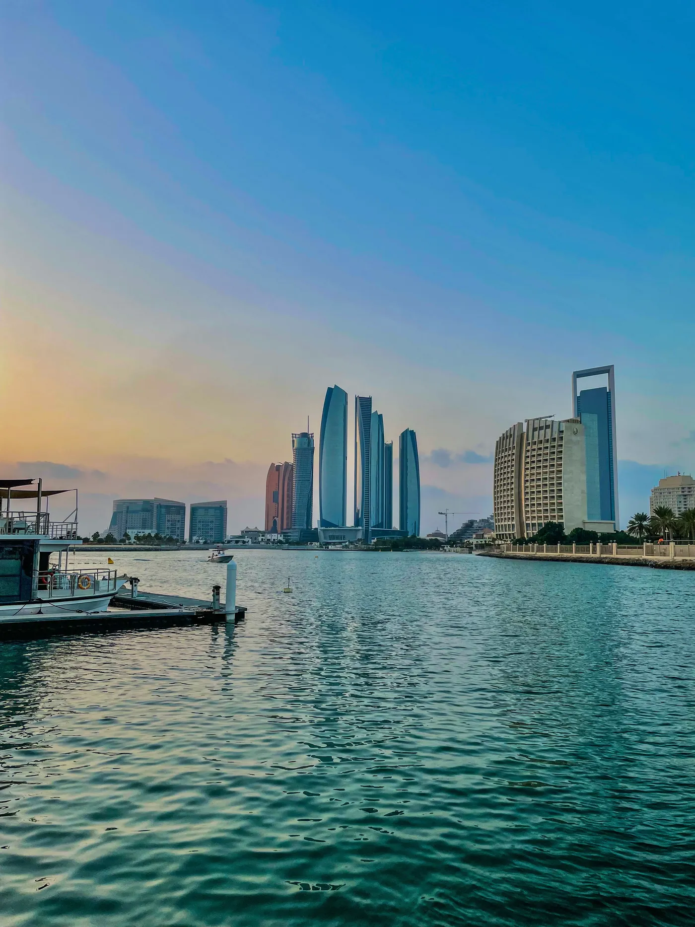 The curved, tapering glass towers of Etihad Towers in Abu Dhabi seen across calm water at sunset, with a jetty in the foreground and the ring-topped ADNOC building to the right