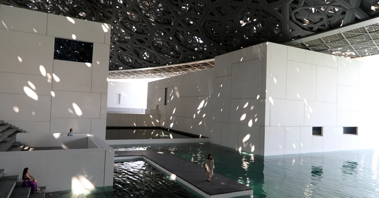 Inside Louvre Abu Dhabi, the perforated dome scattering daylight over white cubic galleries and a channel of seawater with low walkways crossing it