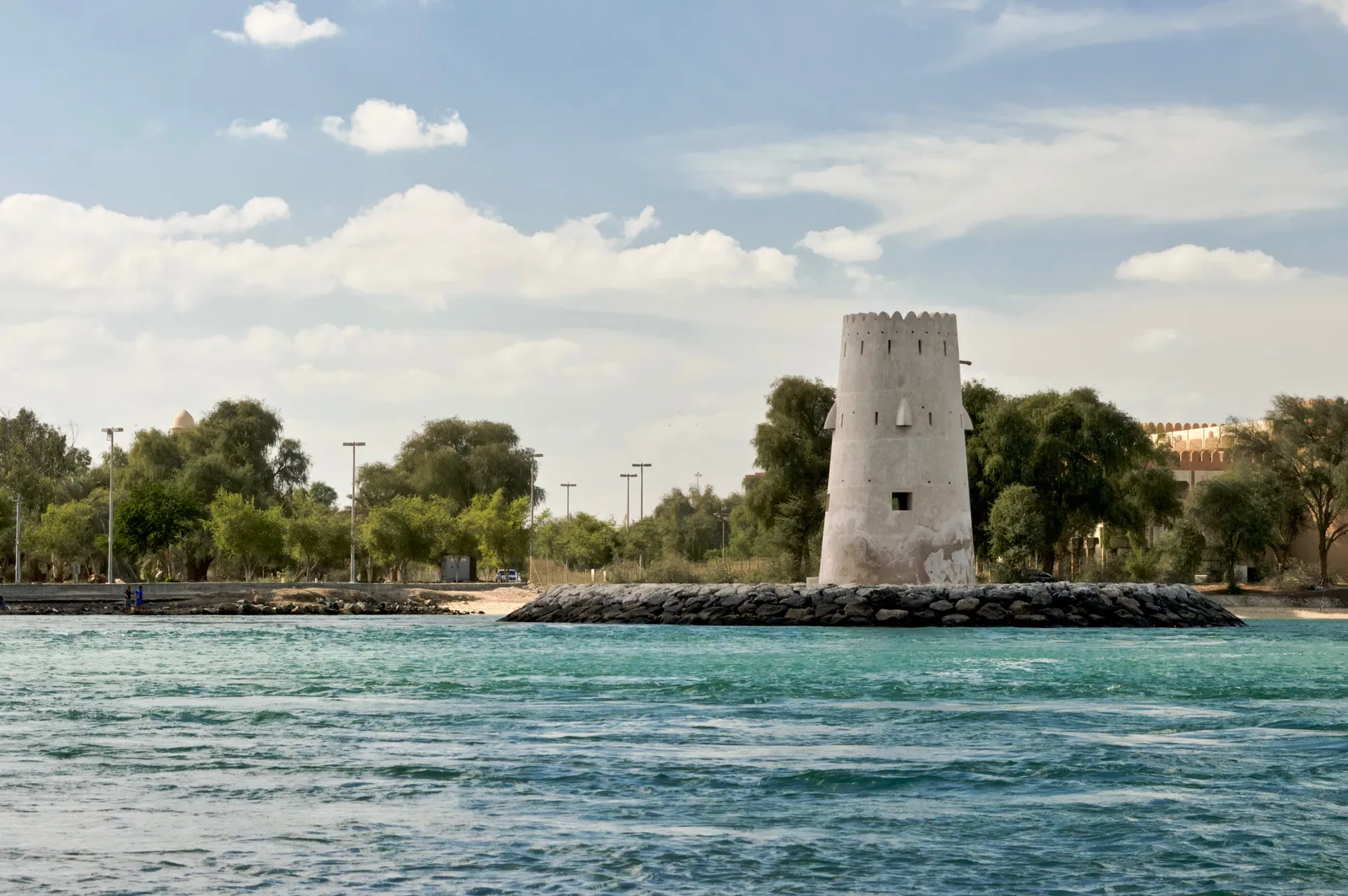 The white crenellated coral-stone watchtower at Al Maqtaa in Abu Dhabi standing on a rocky spit in turquoise water, with trees along the bank behind it