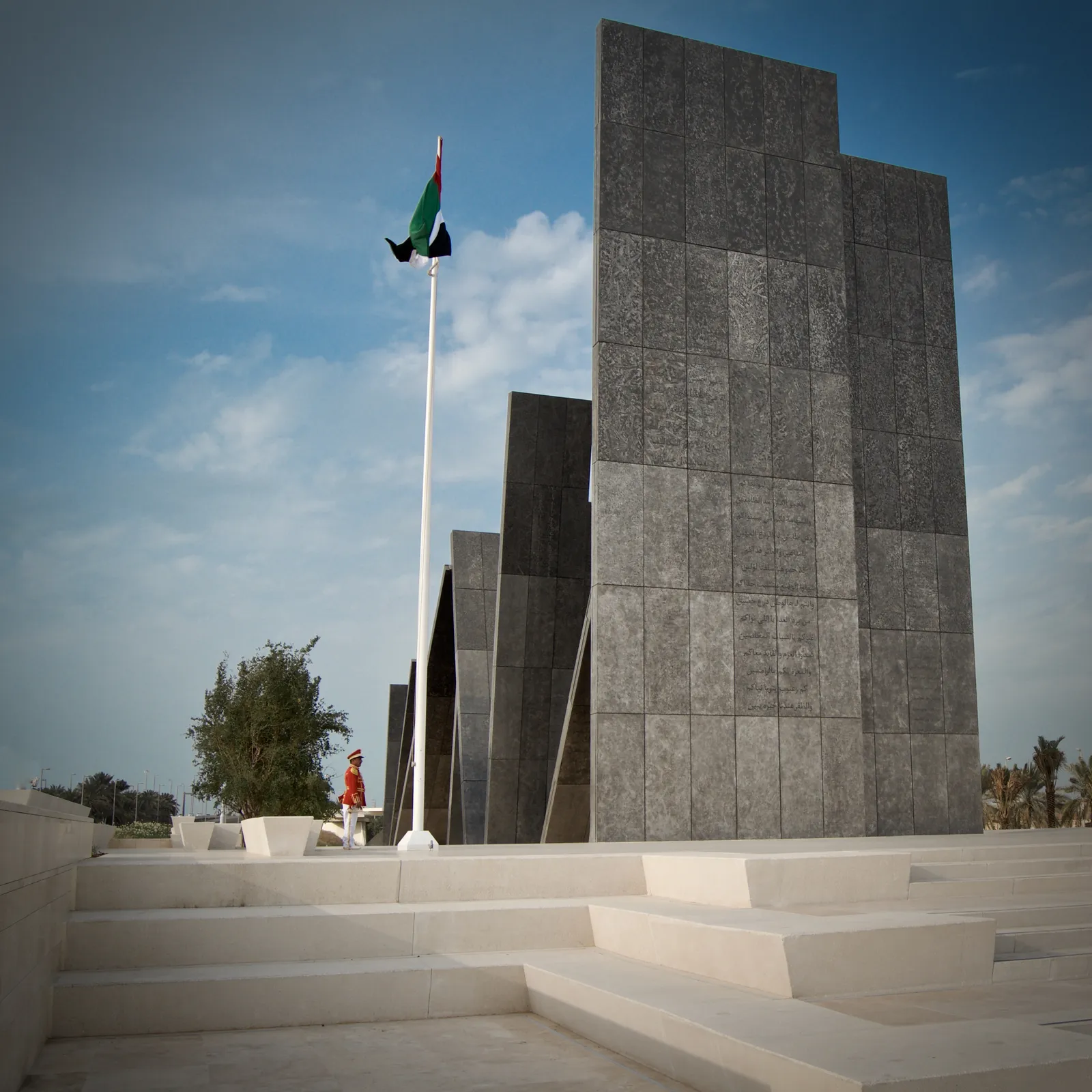 The Wahat Al Karama memorial in Abu Dhabi, tall panelled slabs leaning against one another on a white stone plaza with the UAE flag flying beside them