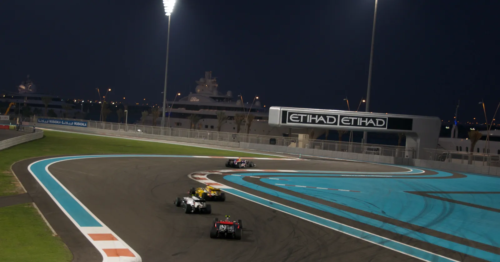Formula 1 cars running under floodlights on the Yas Marina Circuit in Abu Dhabi, with blue-painted run-off areas alongside the track and grandstands behind