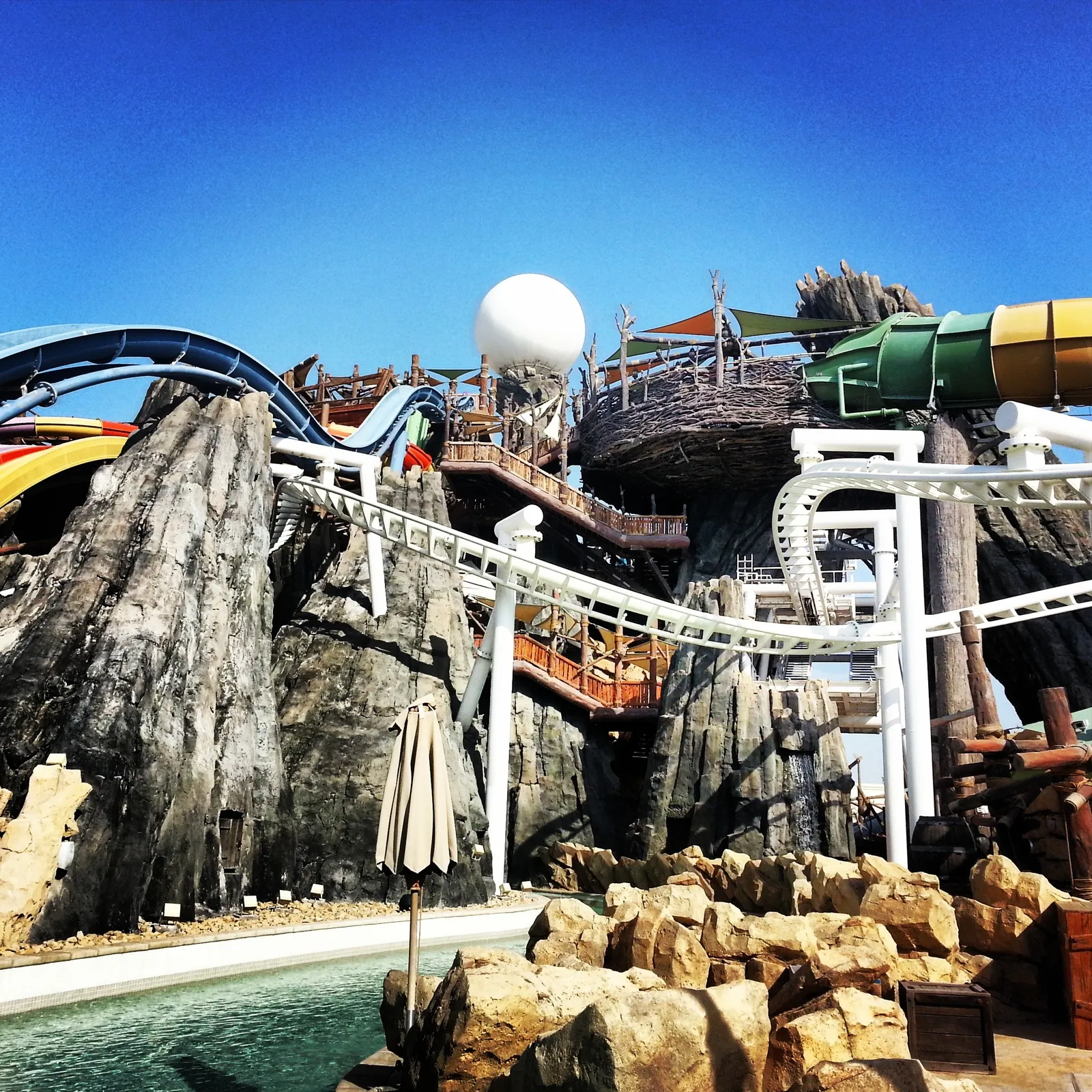 Slides and walkways winding between artificial rock formations at Yas Waterworld in Abu Dhabi, with a large white pearl-shaped structure at the top under a clear blue sky