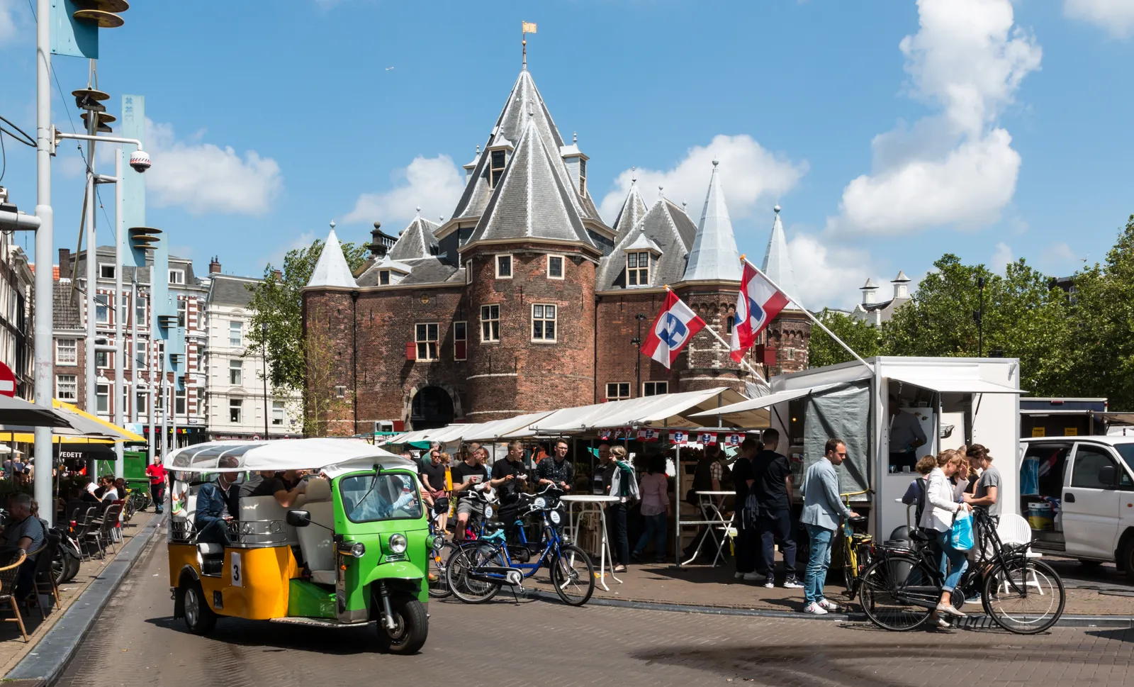 The Waag, a turreted medieval brick building, standing in the middle of Nieuwmarkt square in Amsterdam with market stalls, café awnings and parked bicycles around it