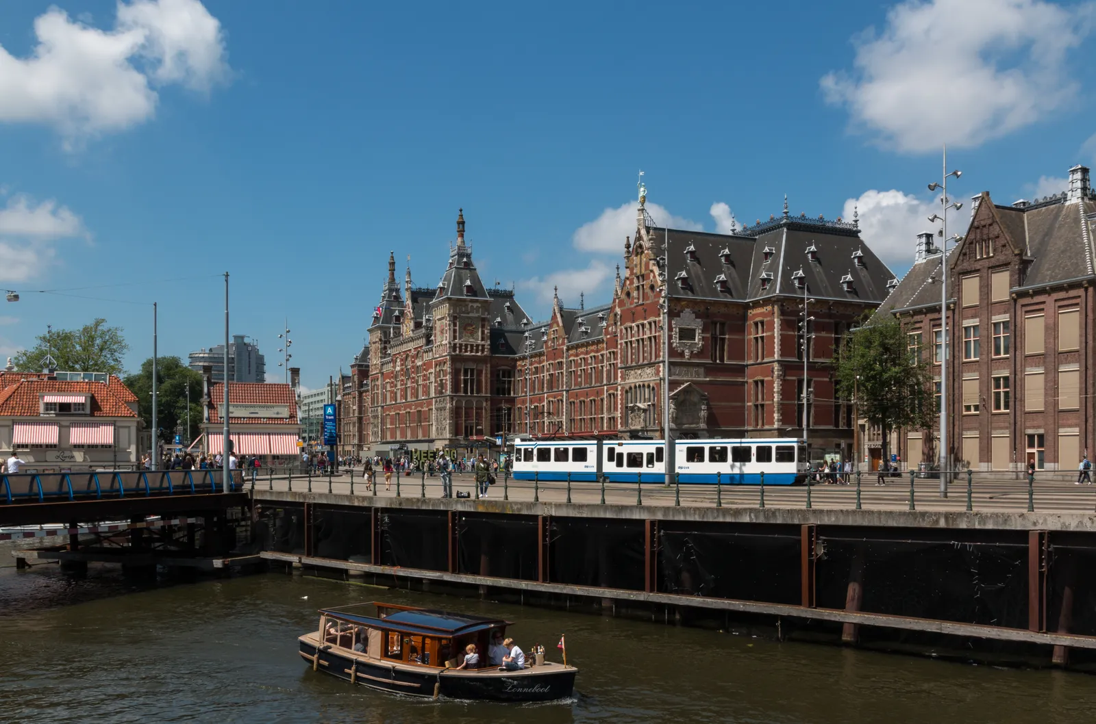 The red brick neo-Renaissance frontage of Amsterdam Centraal station seen across the water on a bright day, with a blue and white GVB tram passing in front and a small canal boat in the foreground