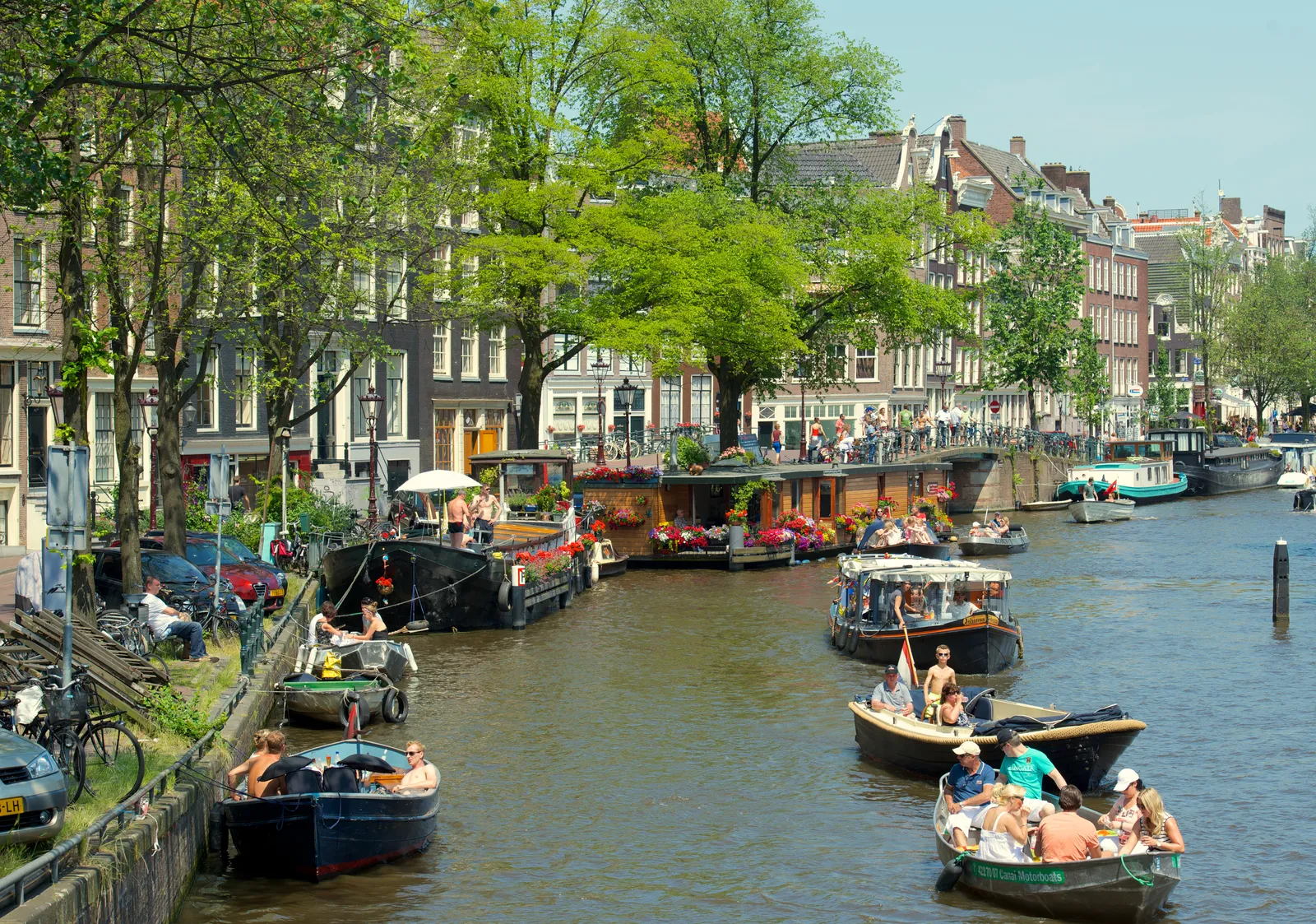 A leafy canal in the Jordaan district of Amsterdam in summer, with flower-covered houseboats moored along the quay and several small open boats full of people on the water