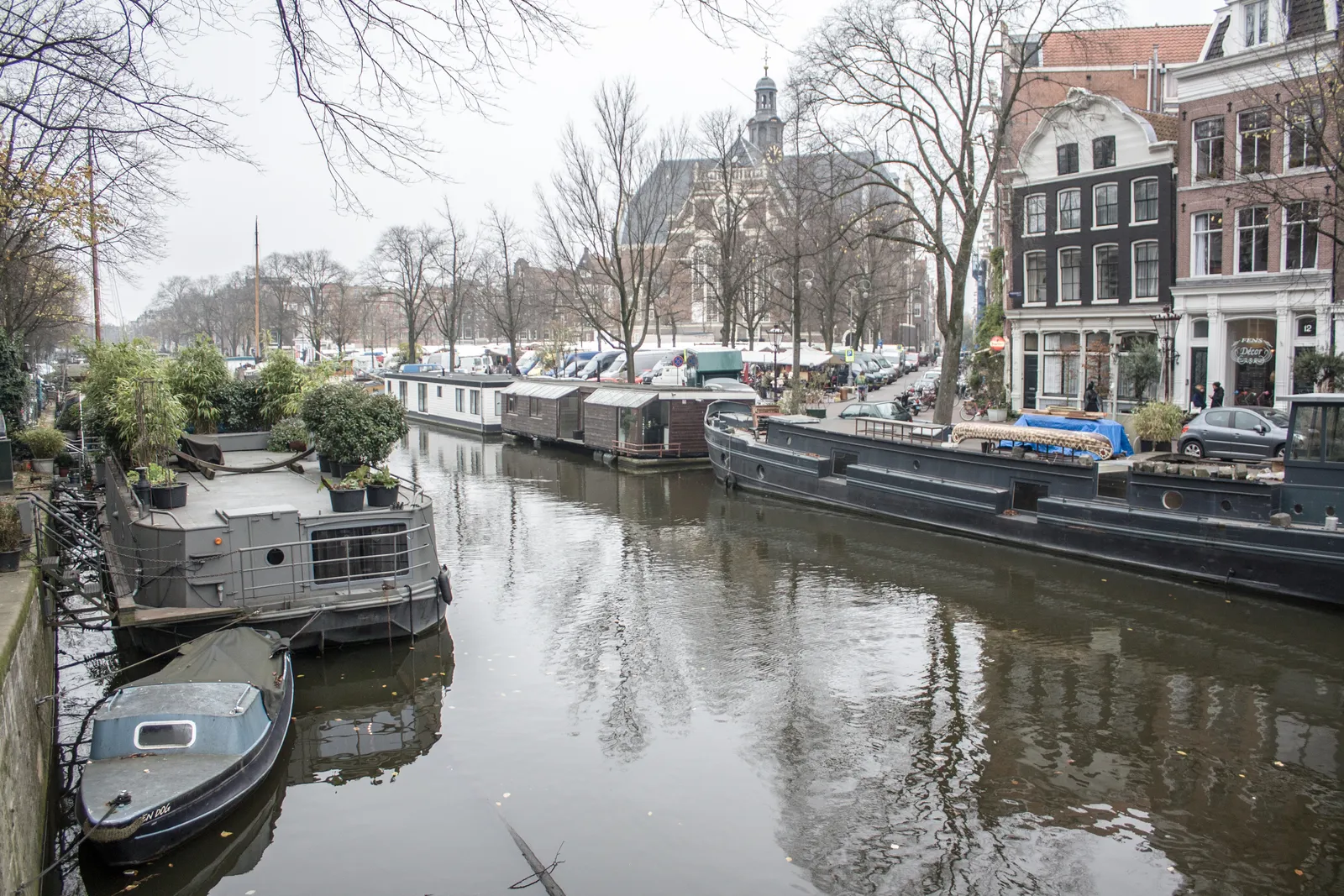 The Prinsengracht canal beside the Noordermarkt in Amsterdam on a grey winter day, lined with houseboats, with market stalls under bare trees and the domed Noorderkerk behind them