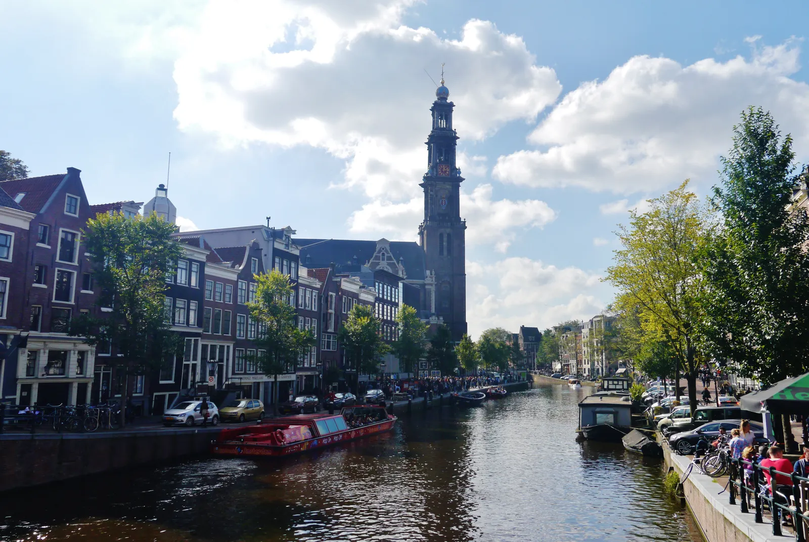 Looking down the Prinsengracht canal in Amsterdam on a bright day, with the tall spire of the Westerkerk tower rising above the gabled houses and a queue of people along the left-hand quay
