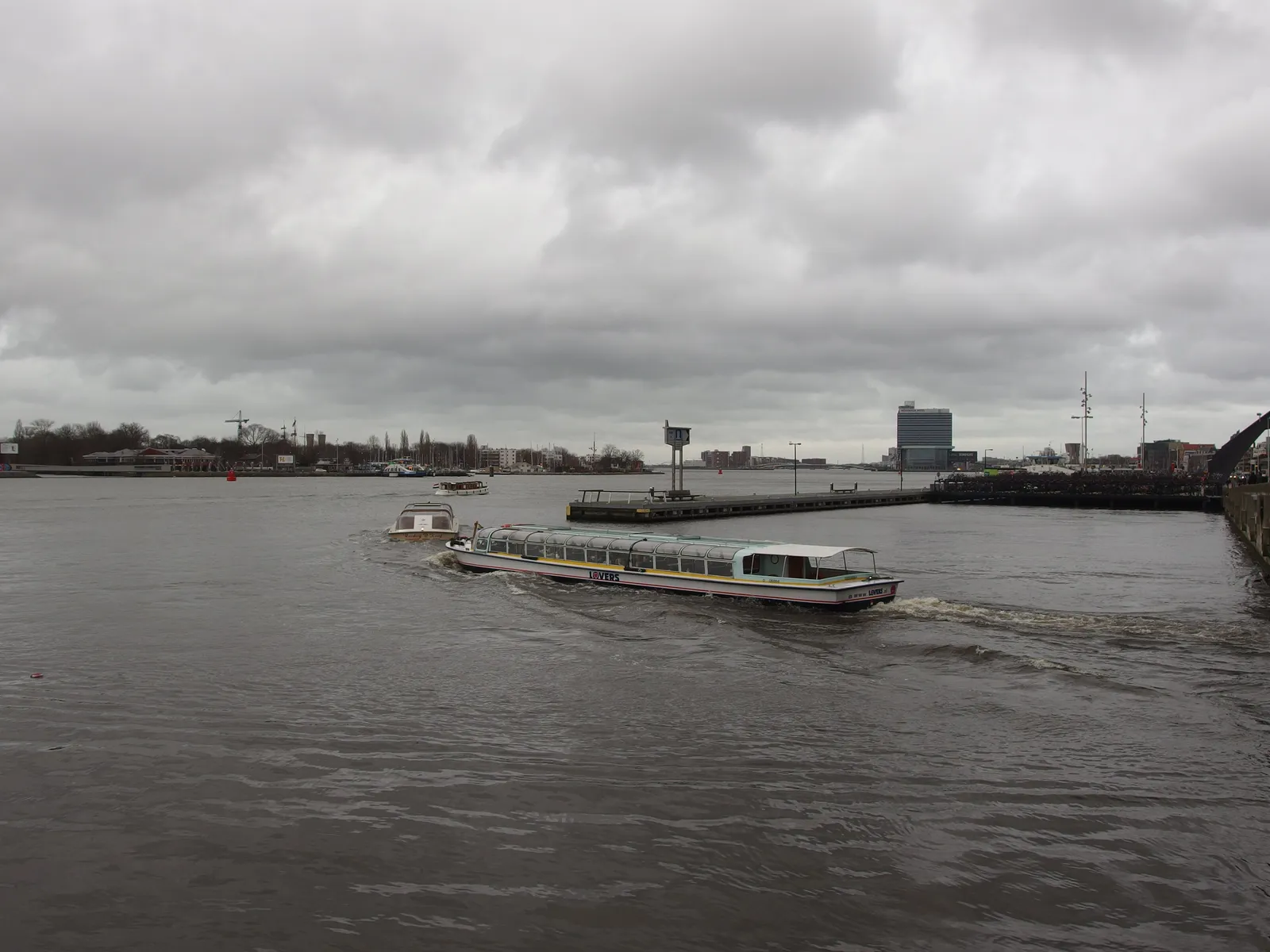 A white and blue passenger ferry crossing the grey water of the IJ in Amsterdam under an overcast sky, with the city skyline visible on the far shore