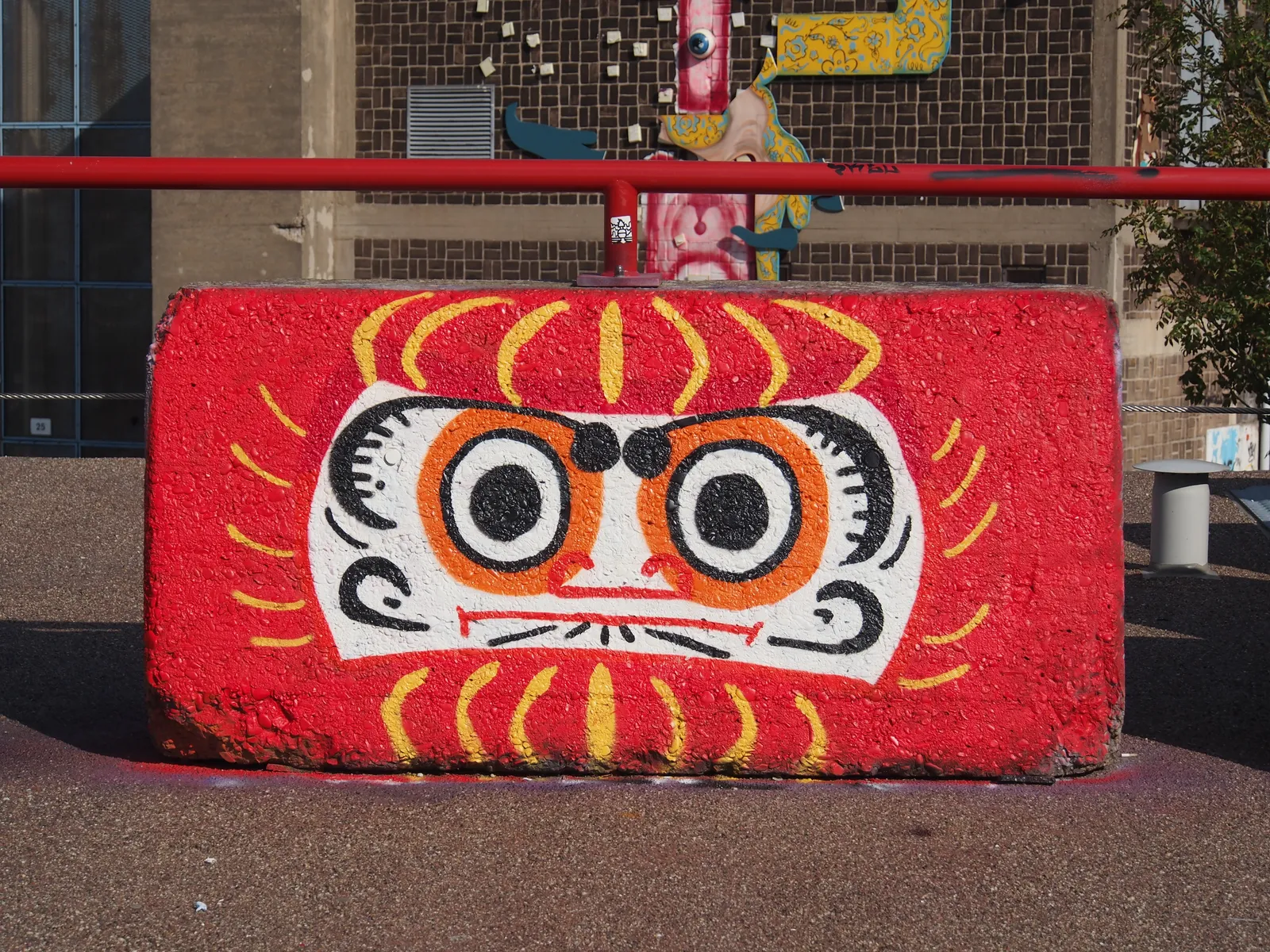 A concrete block in the yard at NDSM painted with a bold red and yellow cartoon face, with a painted brick warehouse wall and mounted sculptural pieces behind it