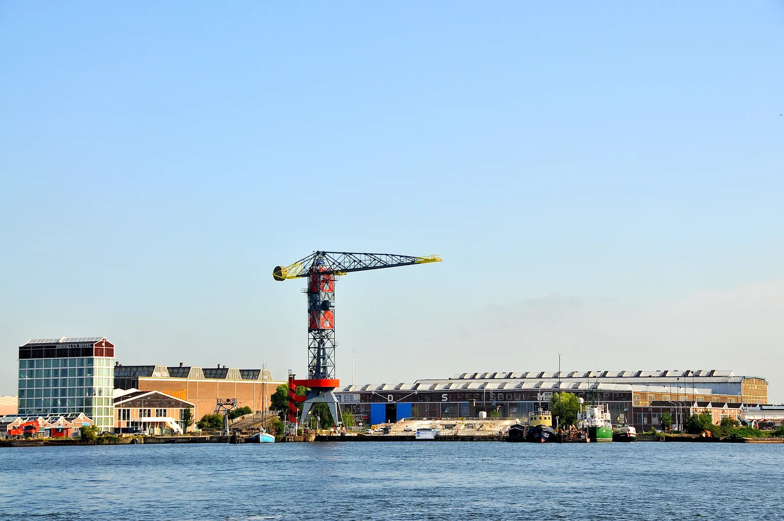 The NDSM shipyard in Amsterdam Noord seen across the water of the IJ, with a red and yellow gantry crane standing over the long steel-roofed shipbuilding sheds along the quay