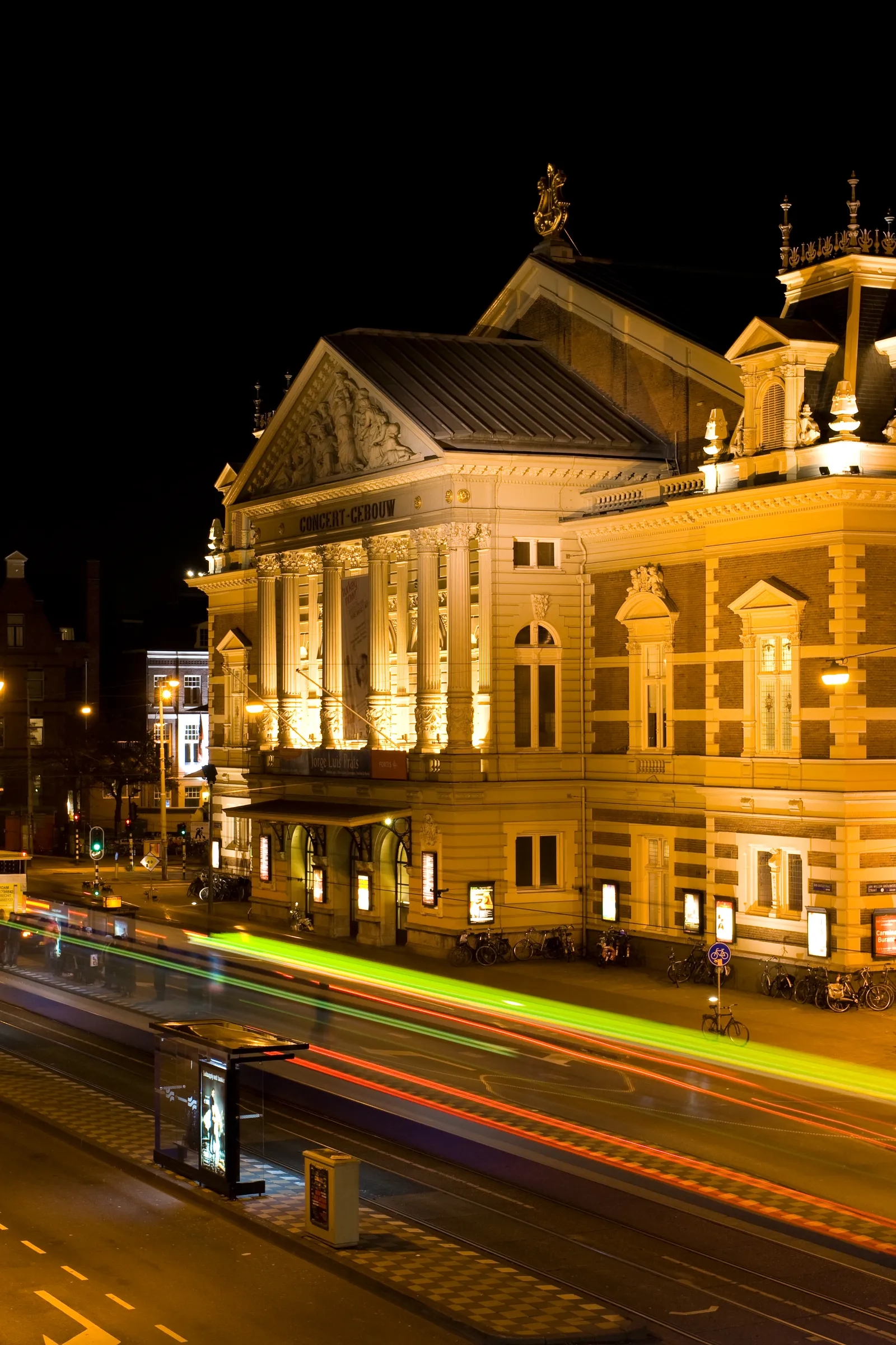 The Concertgebouw concert hall in Amsterdam lit up at night, its classical portico and lettering glowing above the street, with tram light trails streaking across the foreground