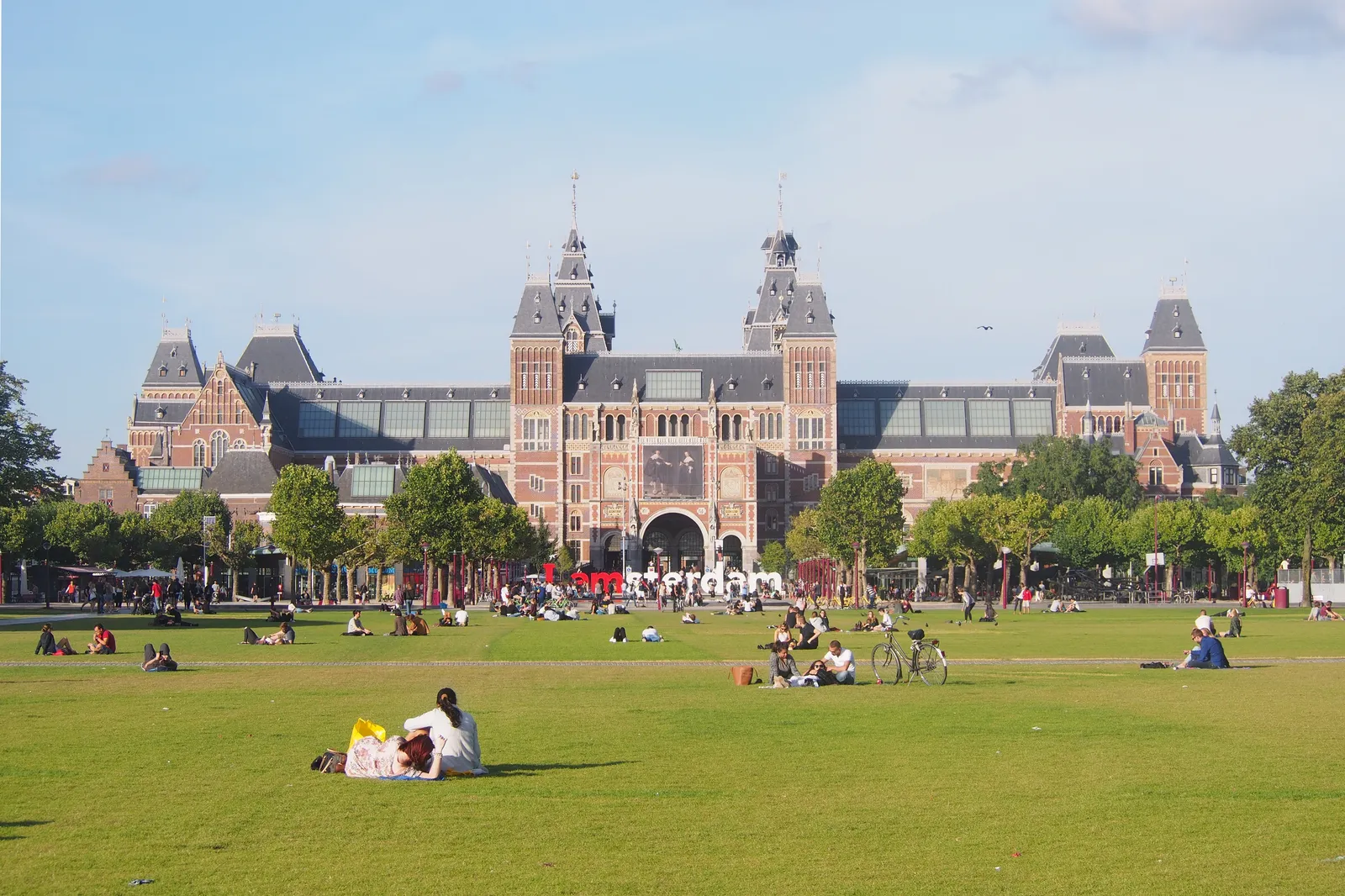 The wide lawn of Museumplein in Amsterdam with people sitting and lying on the grass, and the long brick and stone frontage of the Rijksmuseum across the square behind a line of trees