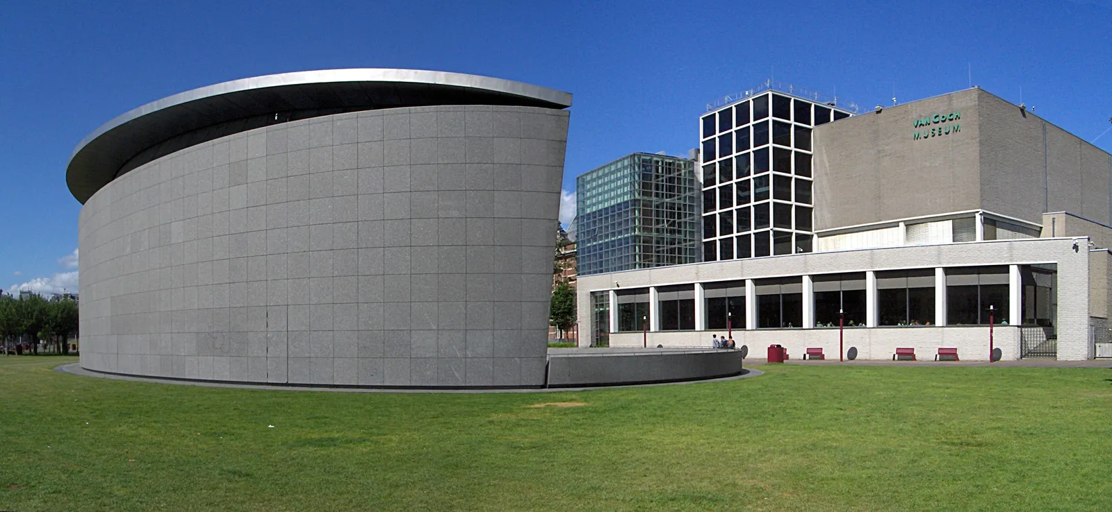 The Van Gogh Museum in Amsterdam seen across grass, with the curved grey stone drum of the Kurokawa exhibition wing in front of the older Rietveld building carrying the museum's name