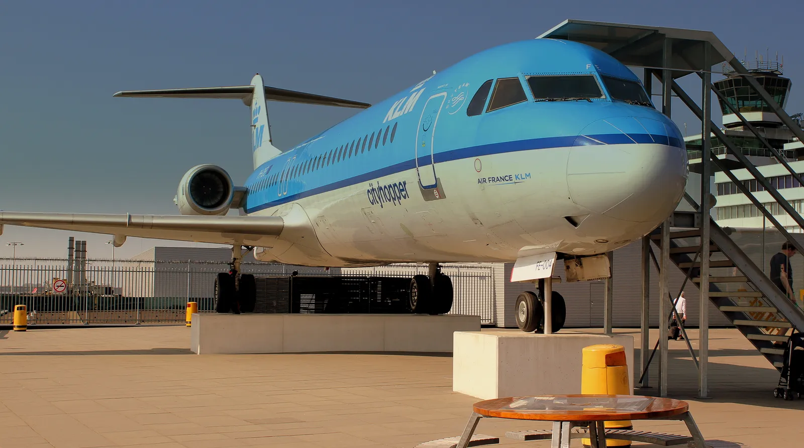 A retired blue and white KLM Cityhopper Fokker 100 parked on the open Panorama Terrace at Schiphol Airport, with the airport's control tower rising behind it