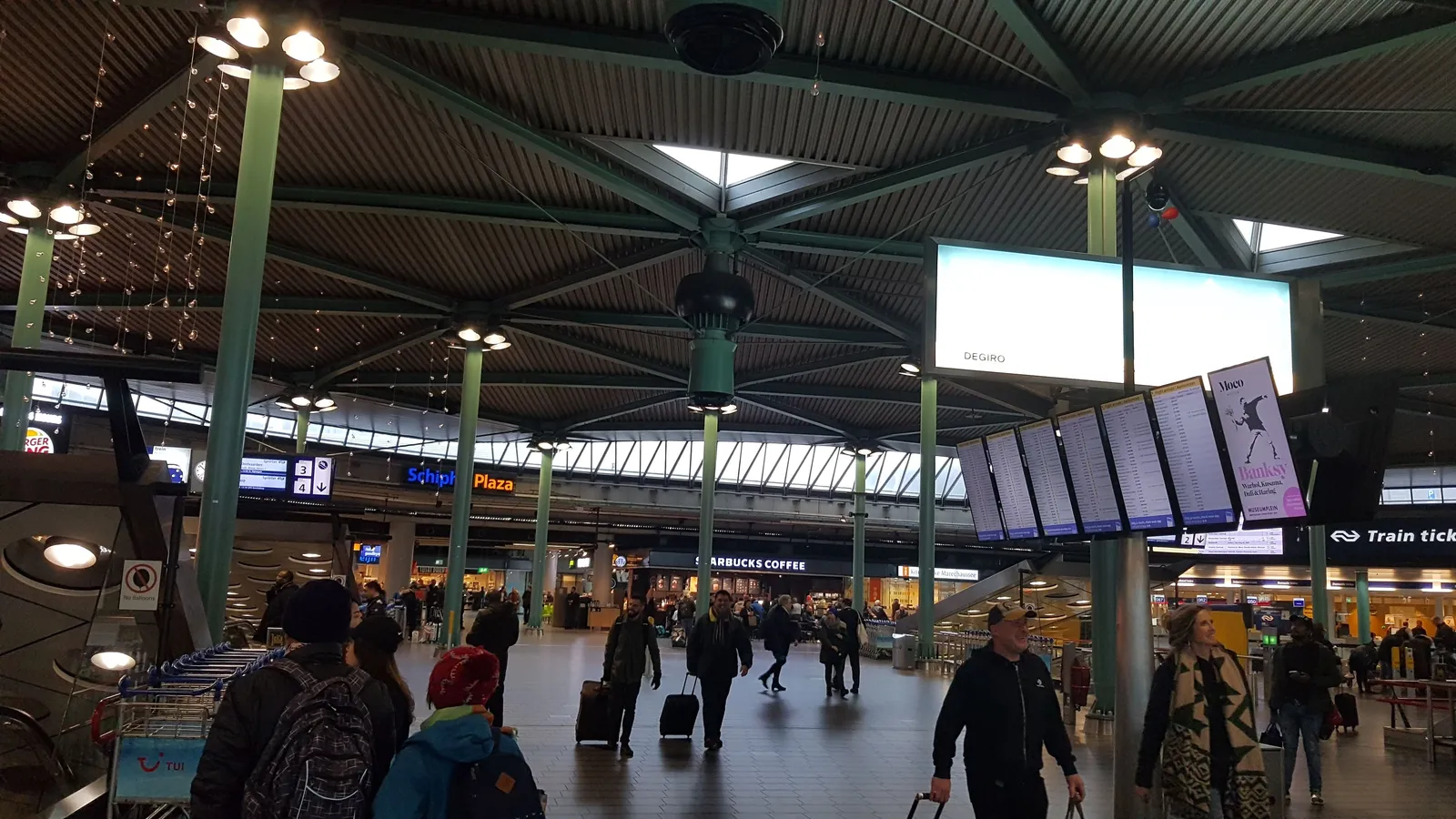 The public concourse of Schiphol Plaza under its tent-like steel roof, with departure boards, a train ticket office and travellers crossing with wheeled cases