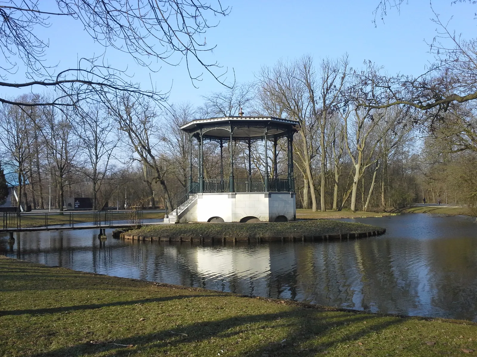 The green cast-iron bandstand in Vondelpark standing on its own small island in the middle of the pond, reached by a footbridge, with bare winter trees behind it under a blue sky