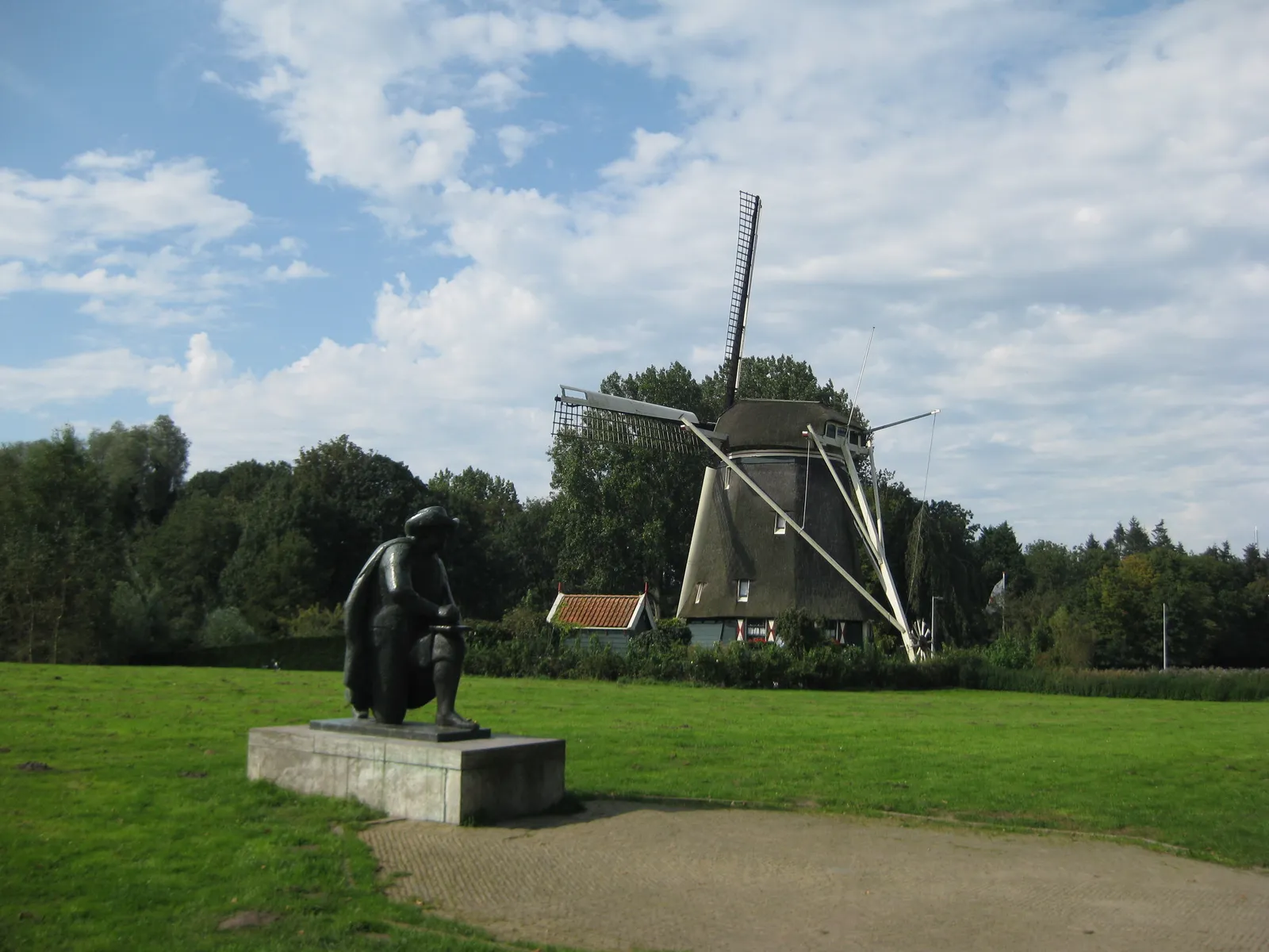The thatched Riekermolen drainage windmill standing on a green riverside meadow beside the Amstel, with a bronze seated statue of Rembrandt sketching in the foreground