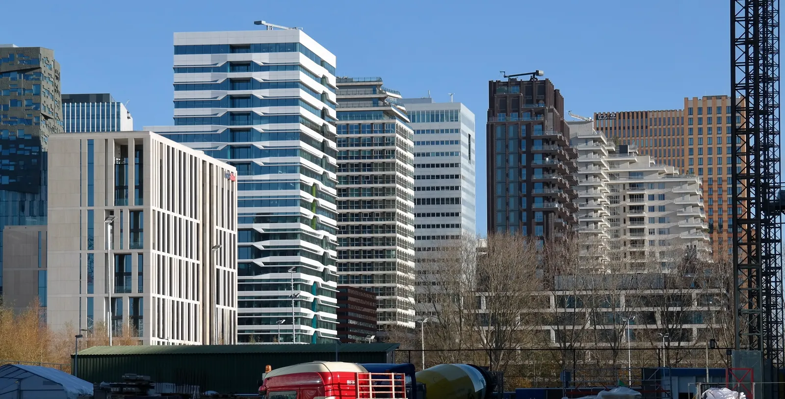 The glass and brick office and apartment towers of the Zuidas business district in Amsterdam under a clear sky, with construction hoardings and a crane at their base