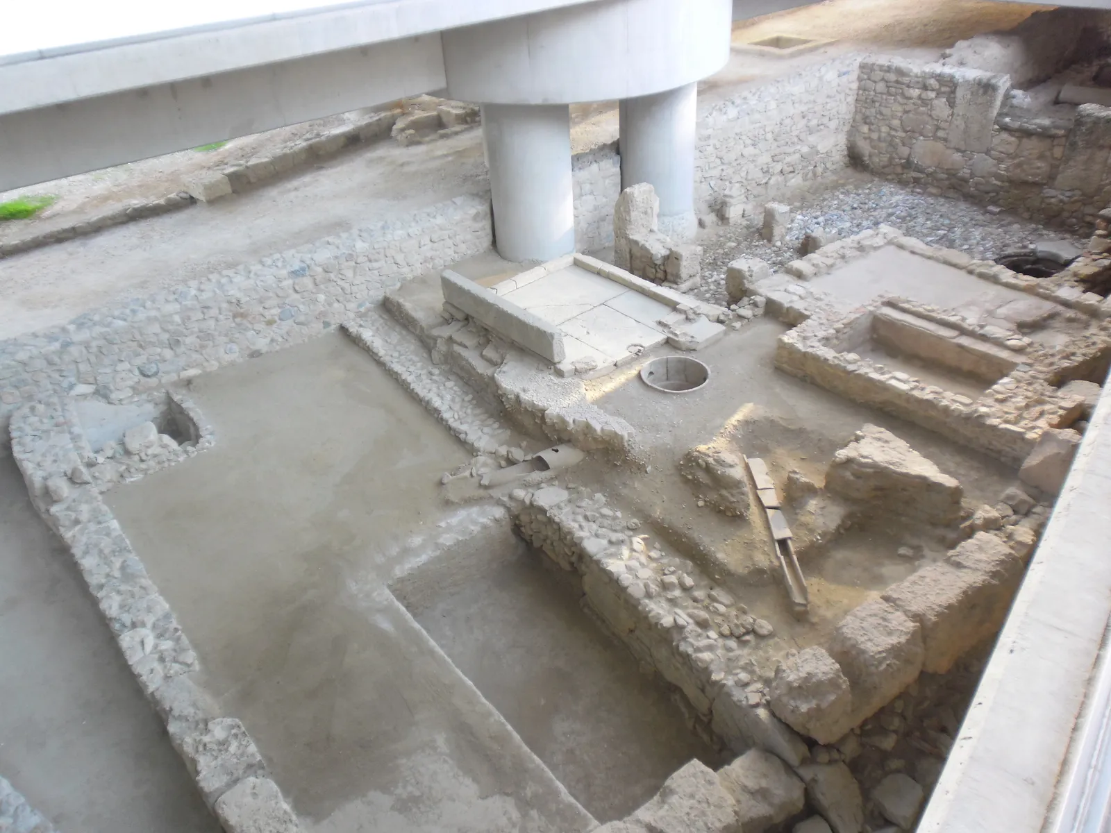 Excavated stone walls, rooms and a mosaic floor beneath the Acropolis Museum in Athens, with one of the building's concrete columns landing between them