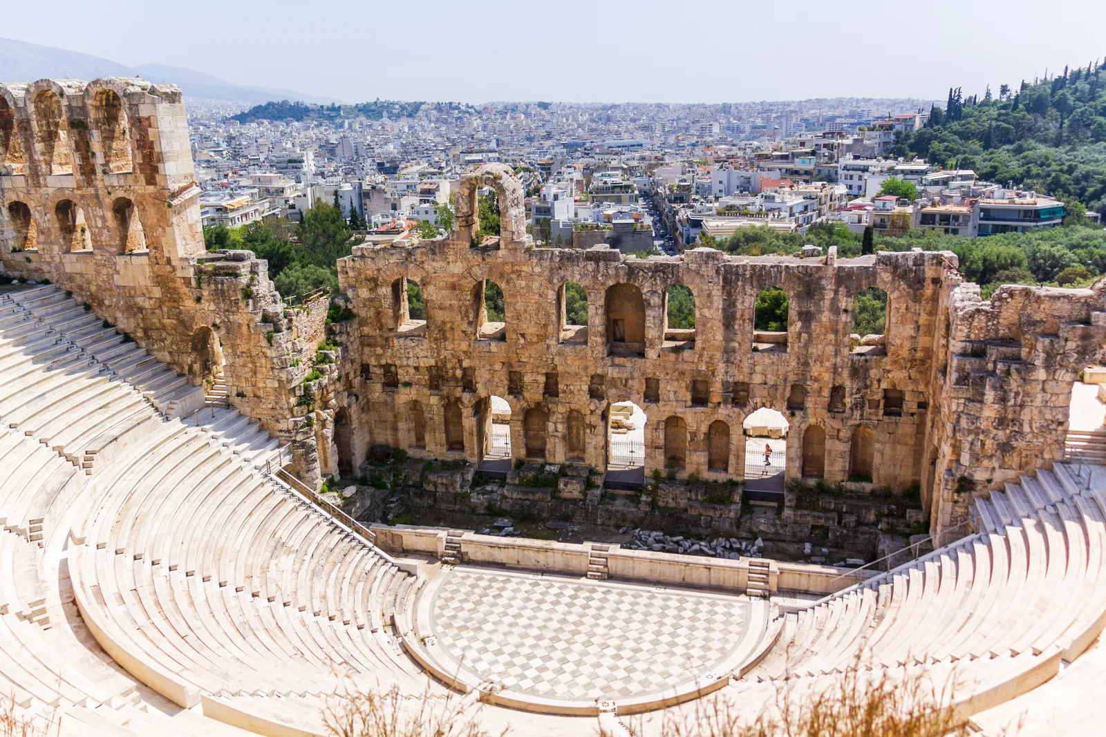 The Odeon of Herodes Atticus in Athens seen from above, its curved tiers of stone seating facing a tall arched stage wall with the city behind