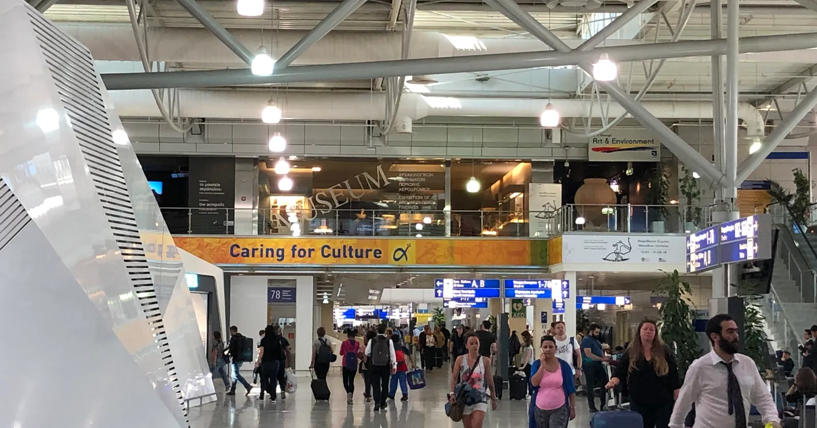 The main concourse of Athens International Airport with travellers wheeling suitcases below the raised mezzanine that holds the airport's museum