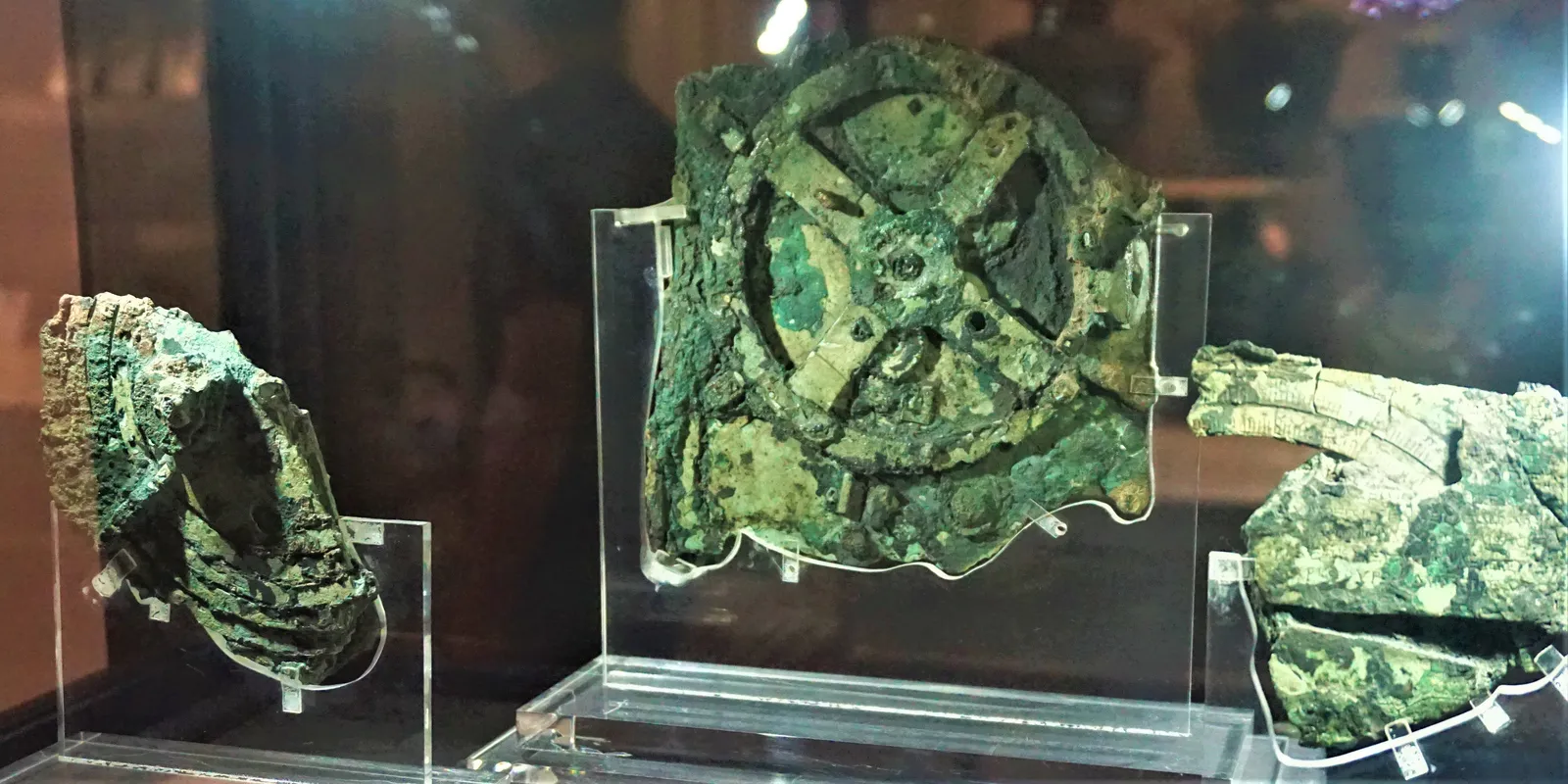 Three corroded green bronze fragments of the Antikythera Mechanism in a display case, the largest showing a toothed gear wheel