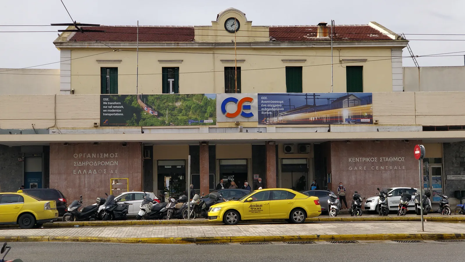 The low ochre street front of Athens Larissa railway station with a clock above the entrance and taxis waiting outside
