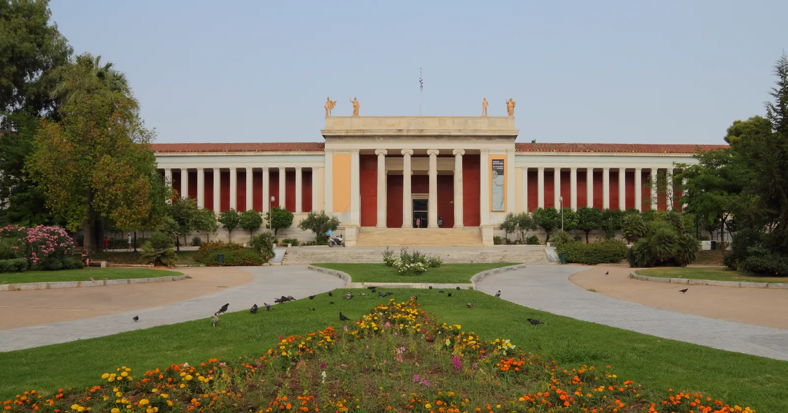 The long neoclassical facade of the National Archaeological Museum in Athens behind its garden of flowerbeds and gravel paths
