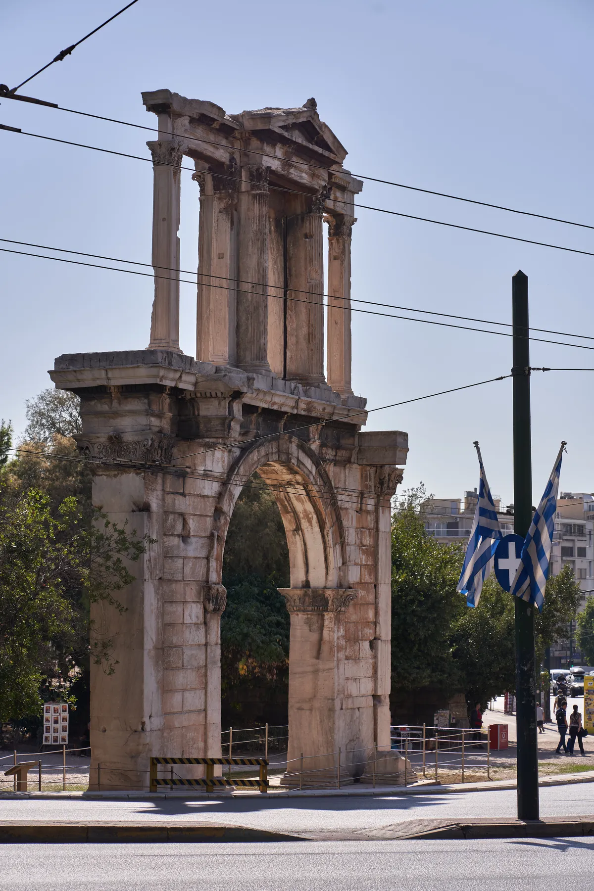 The Arch of Hadrian in Athens standing directly beside the roadside pavement, with tram wires crossing in front of it and Greek flags to the right