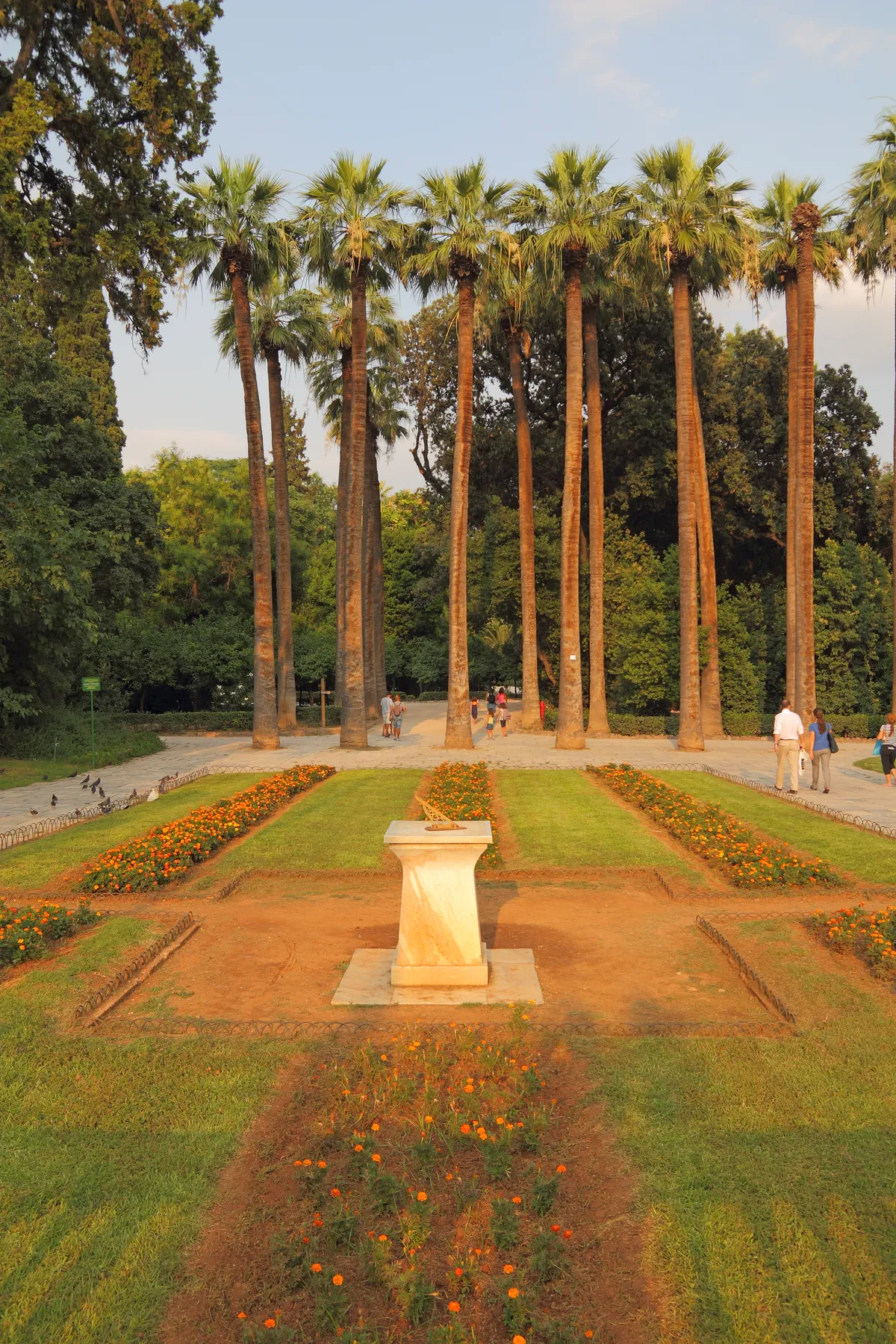 An avenue of tall palms in the National Garden in Athens above formal beds of orange flowers and a marble sundial