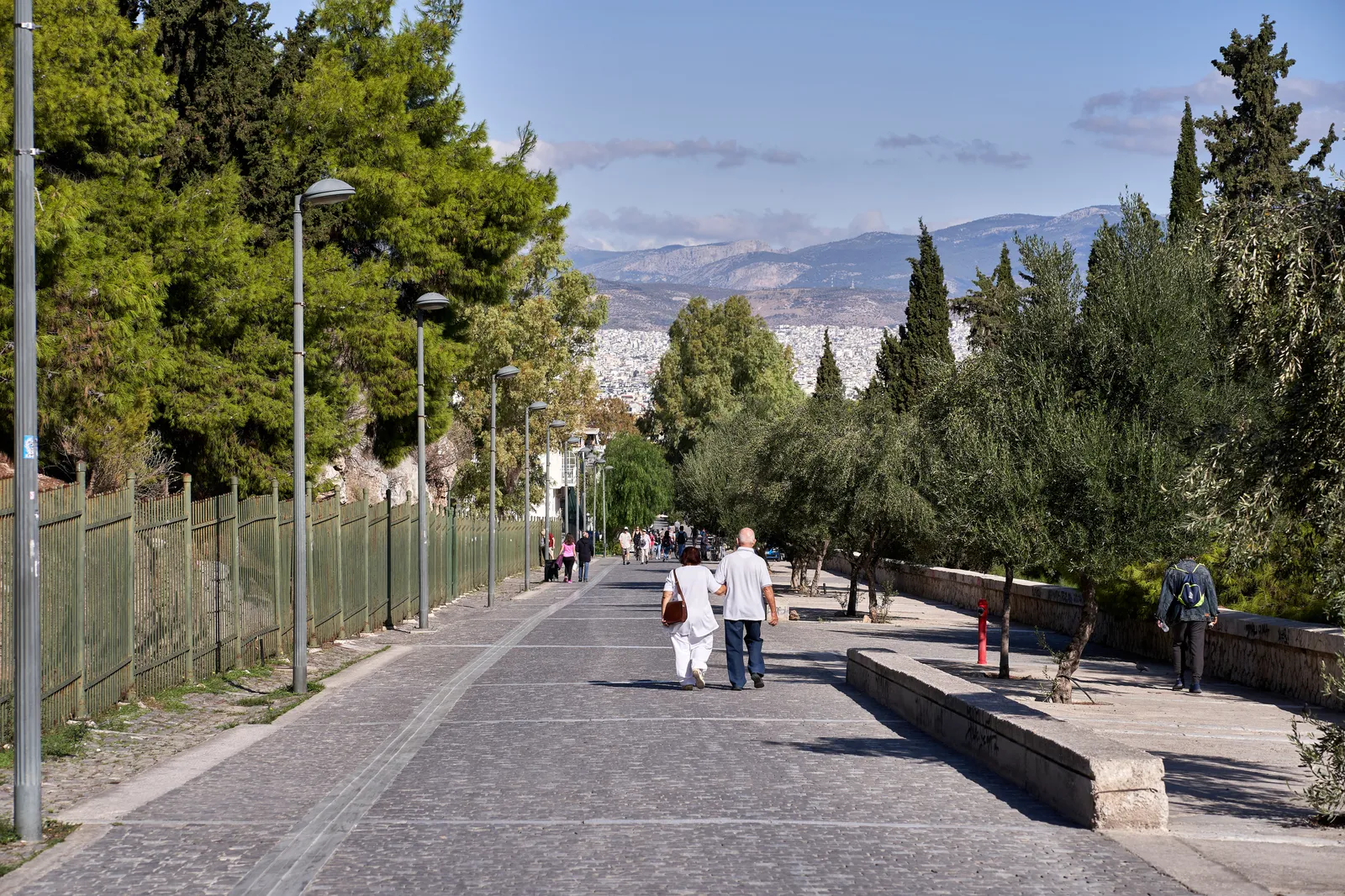 The wide paved Apostolou Pavlou pedestrian street in Athens running between pines and olive trees with walkers on it and the hills of Attica beyond