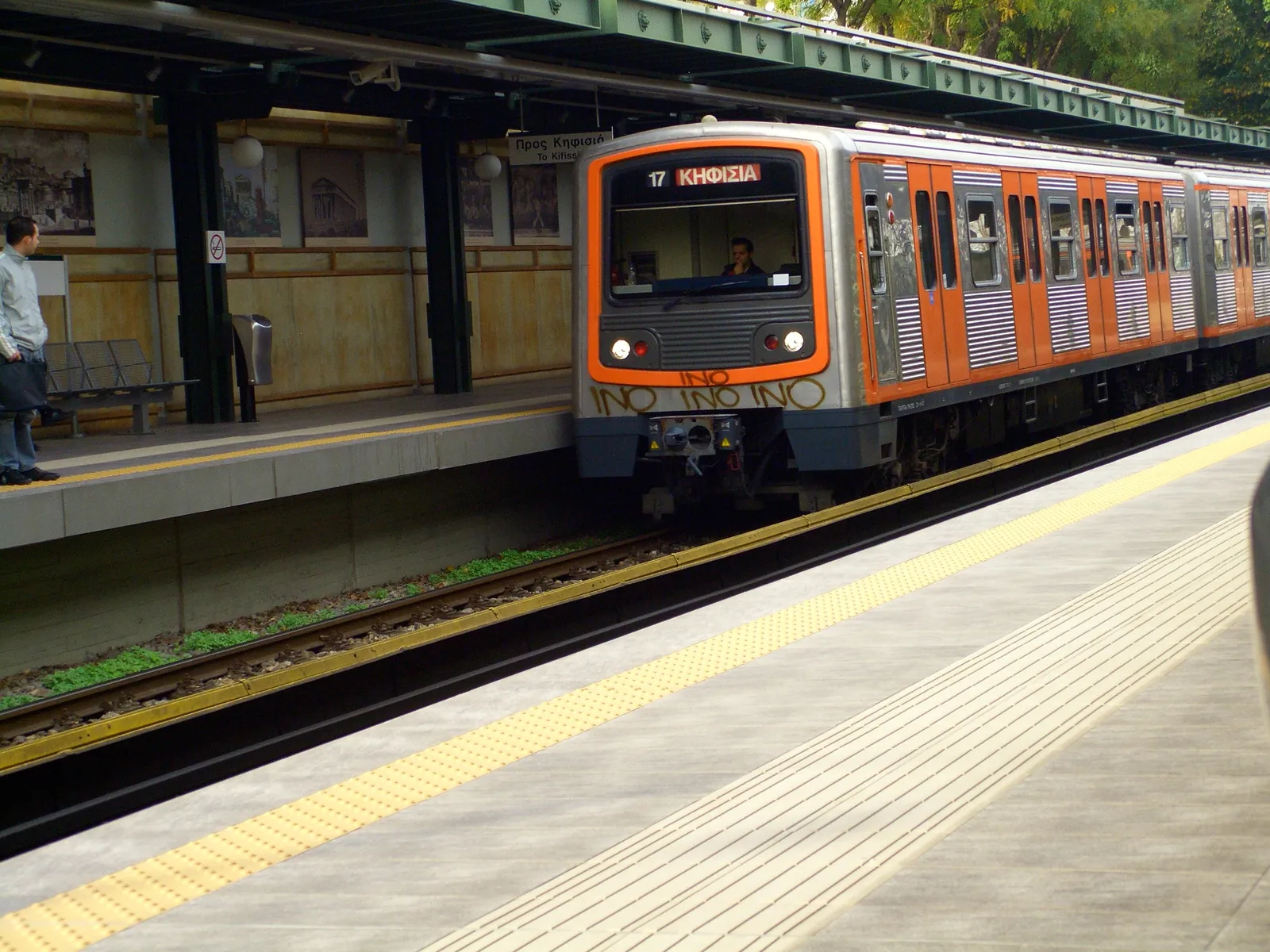 An orange and white Line 1 train pulling into the open-air platform at Thissio station in Athens