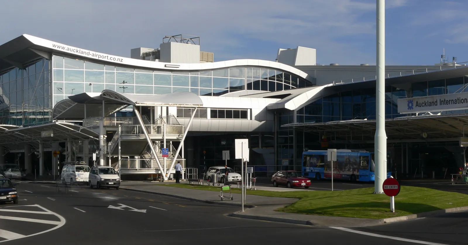 The kerbside frontage of Auckland Airport's international terminal, a long glazed building under a curved white canopy with cars and a shuttle bus on the road in front