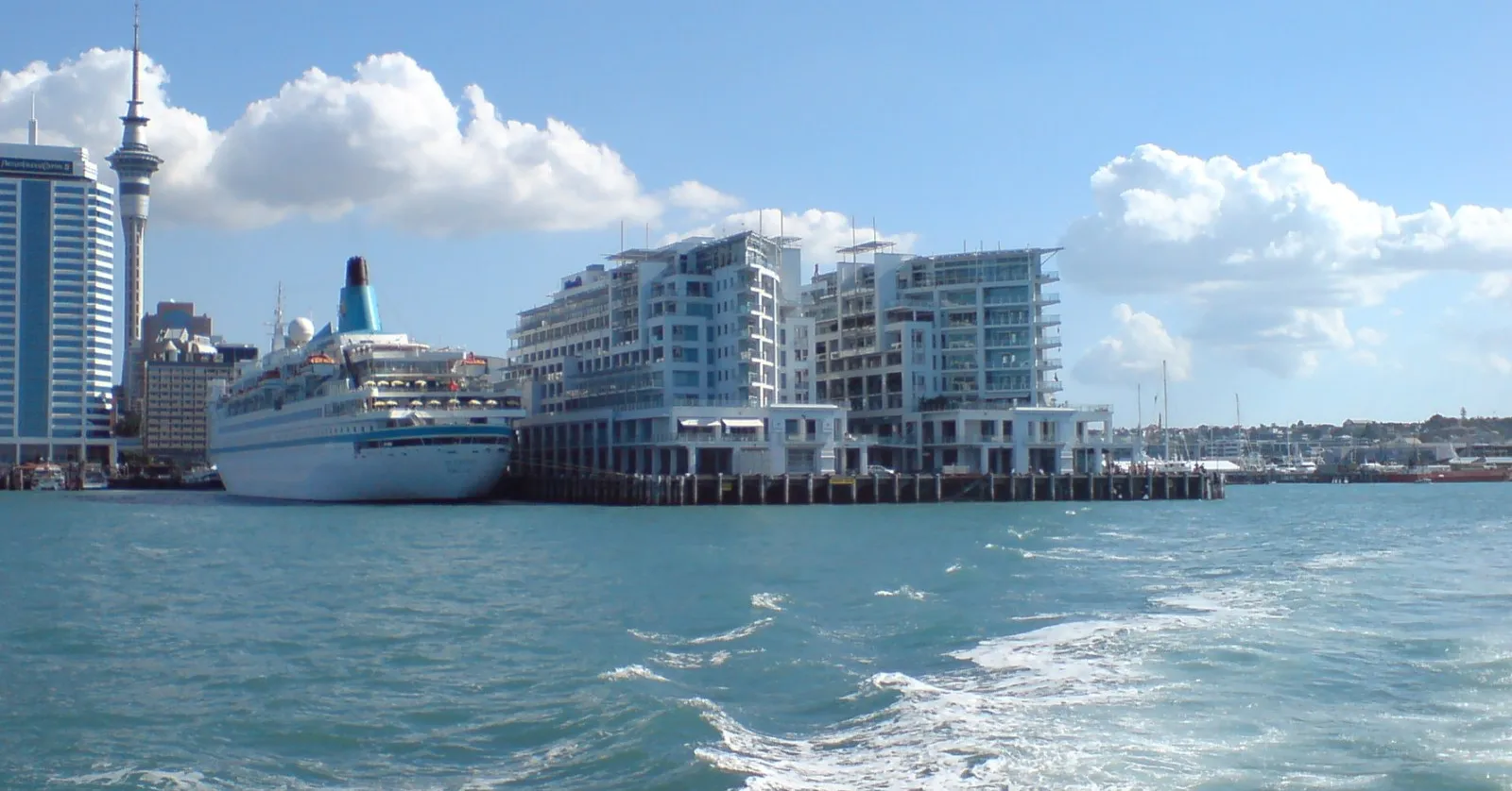 A white cruise ship tied up at Princes Wharf in Auckland, seen from the harbor, with the Sky Tower and the city center's towers behind