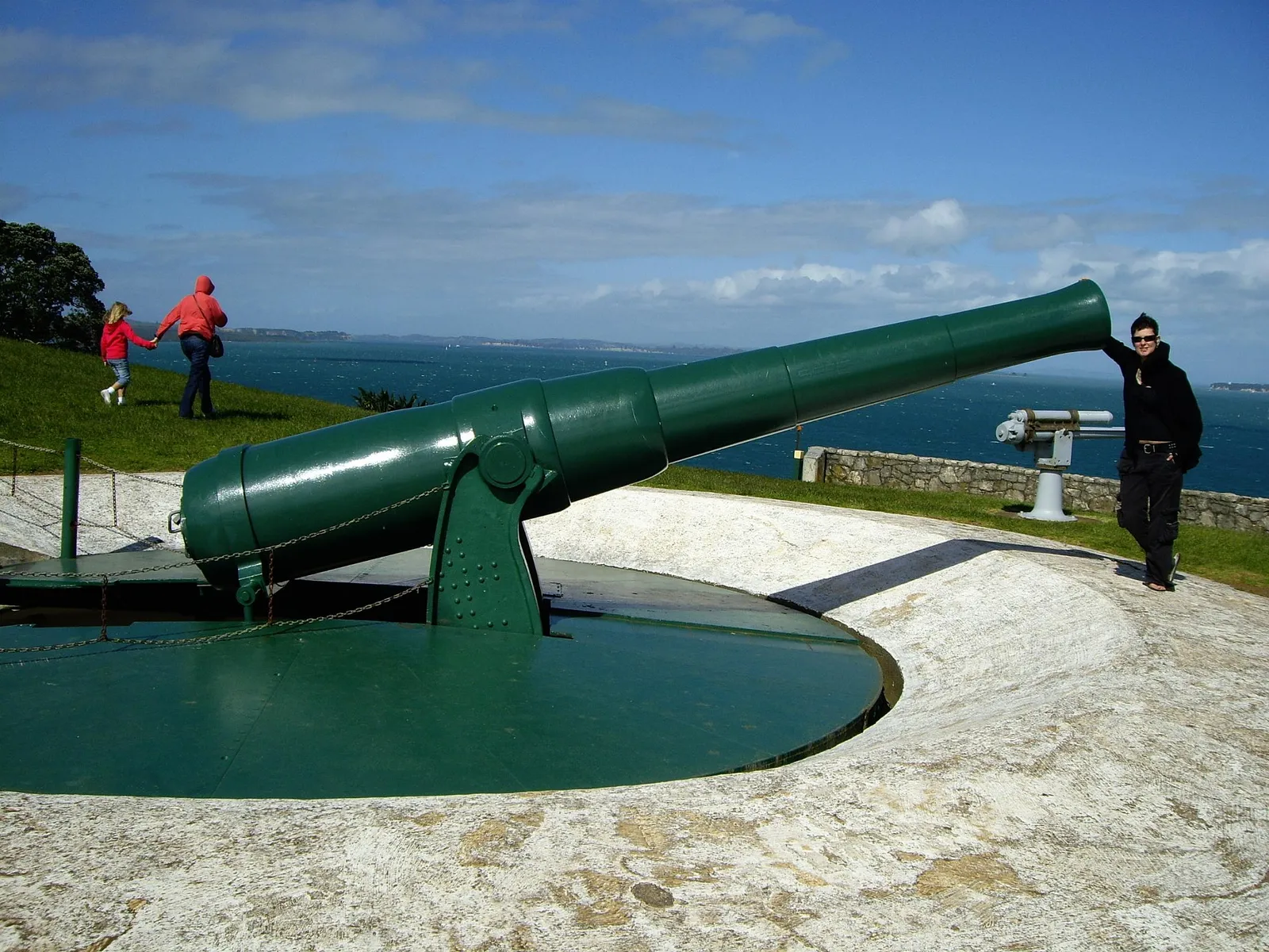 A large green naval gun mounted on a turntable in a circular concrete pit at North Head, Devonport, with the harbour and a grassy headland behind it