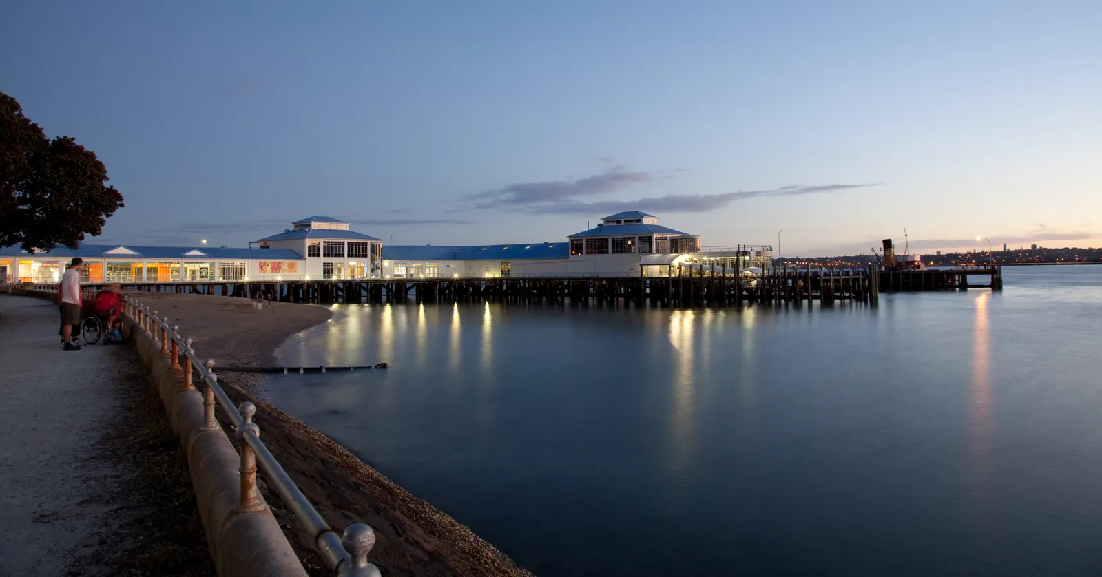 Devonport Ferry Terminal lit at dusk on its timber piles, its canopies reflected in still harbour water, with a seawall and promenade railing in the foreground