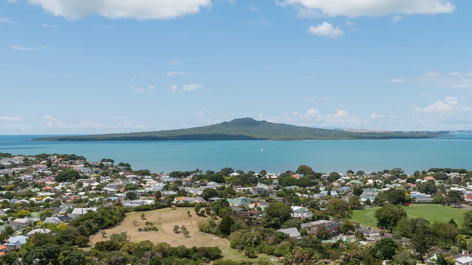 The view from the summit of Mount Victoria in Devonport across rooftops and trees to blue harbour water, with the low cone of Rangitoto Island on the horizon