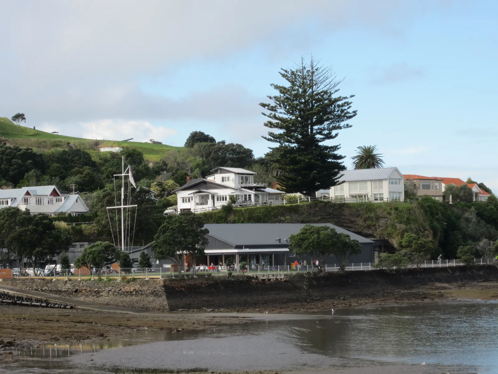 The white heritage buildings of the Torpedo Bay Navy Museum seen from across the bay at low tide, a tall Norfolk pine behind them and a mast standing in the grounds