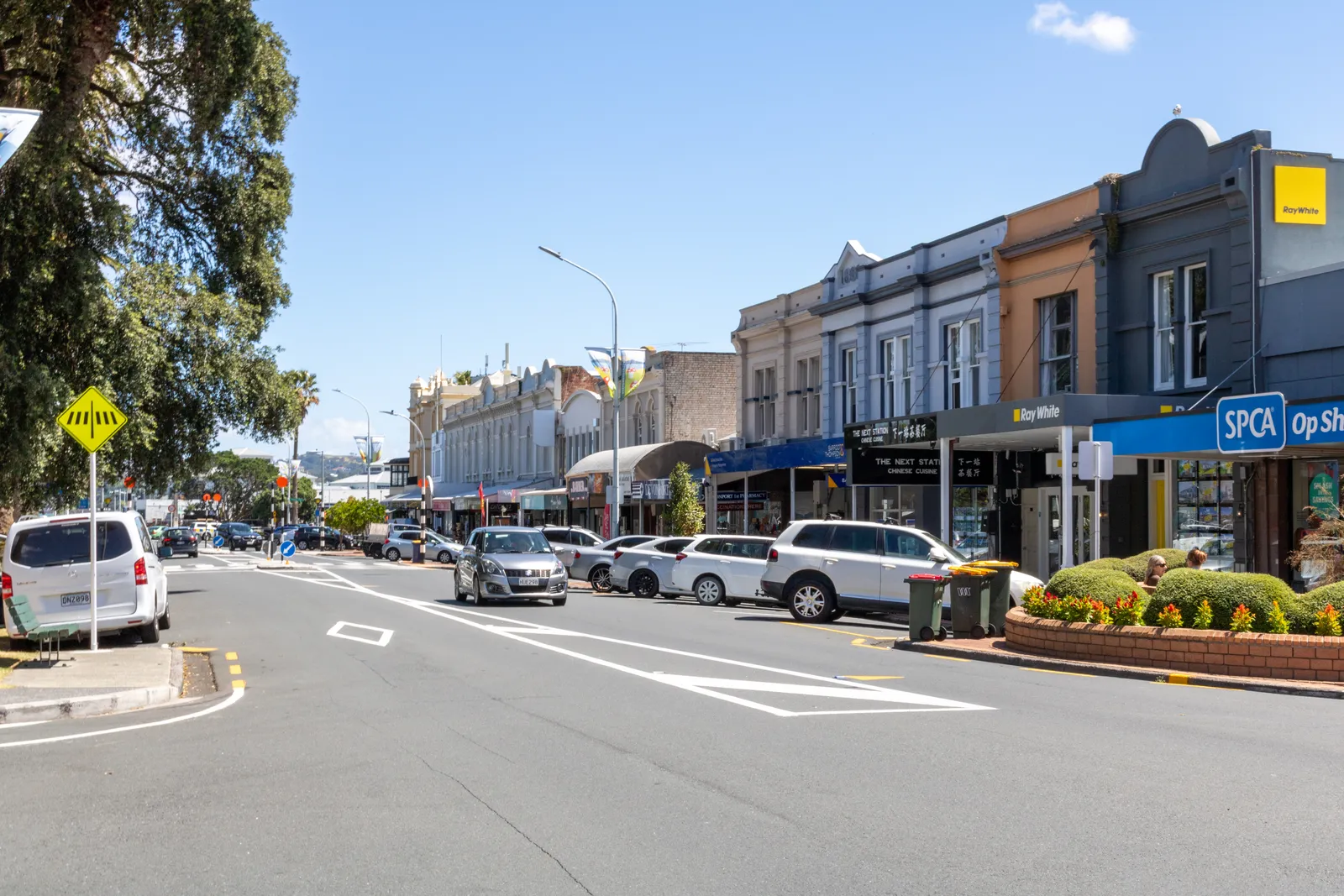 Victoria Road in Devonport on a clear day, a row of Victorian and Edwardian shopfronts with decorated parapets along one side and parked cars down the middle of the street