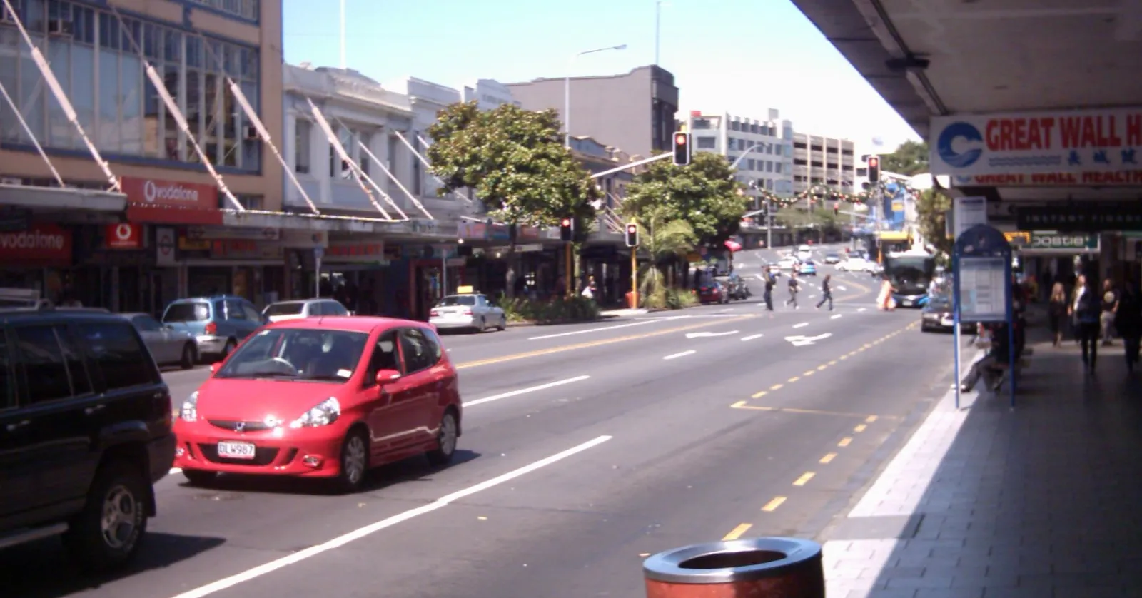 Karangahape Road in Auckland on a clear day, two-storey shopfronts with verandahs along both sides, traffic in the middle and people crossing at the lights further up