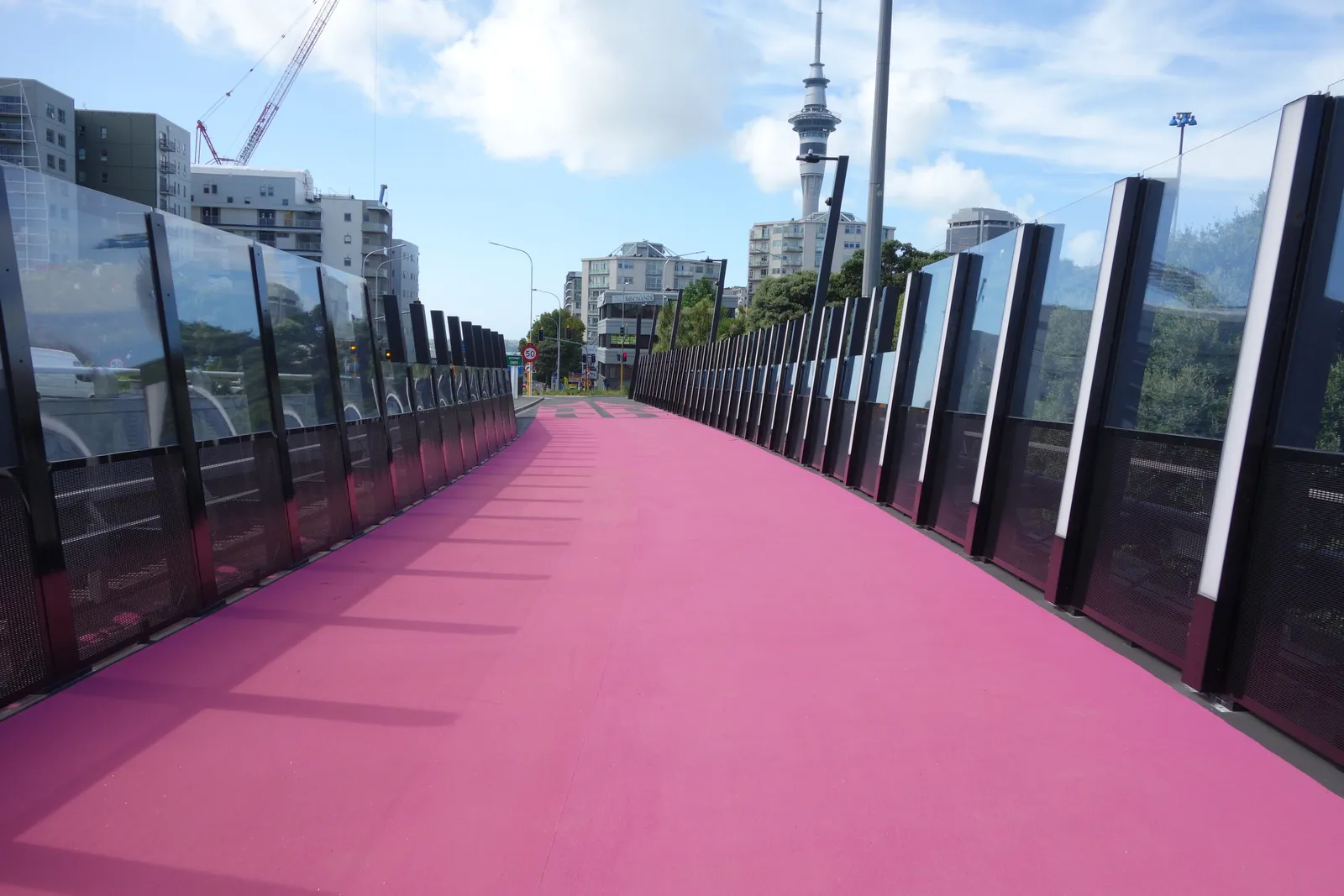 The magenta surface of Te Ara I Whiti curving away between black balustrades lined with lighting poles, apartment blocks and the Sky Tower visible in the distance