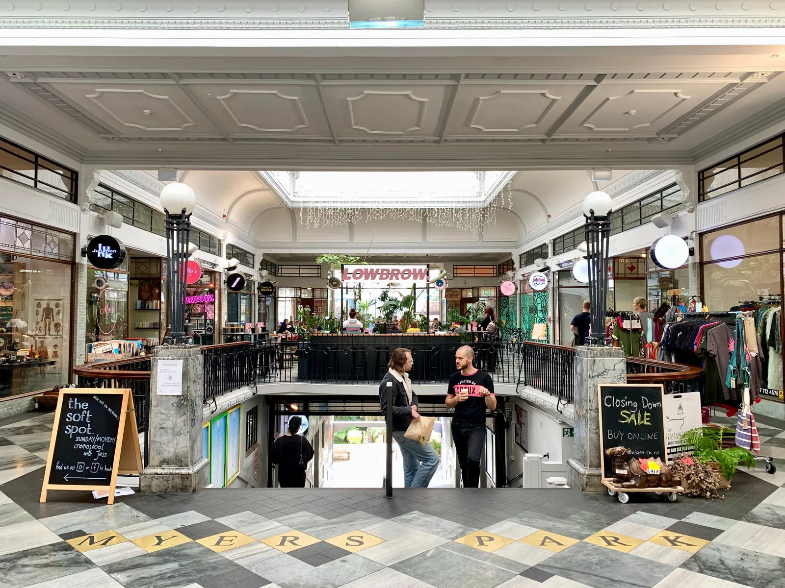 Inside St Kevin's Arcade in Auckland, a pressed plaster ceiling and arched opening above shopfronts, with the words Myers Park set into the tiled floor at the head of a stairway