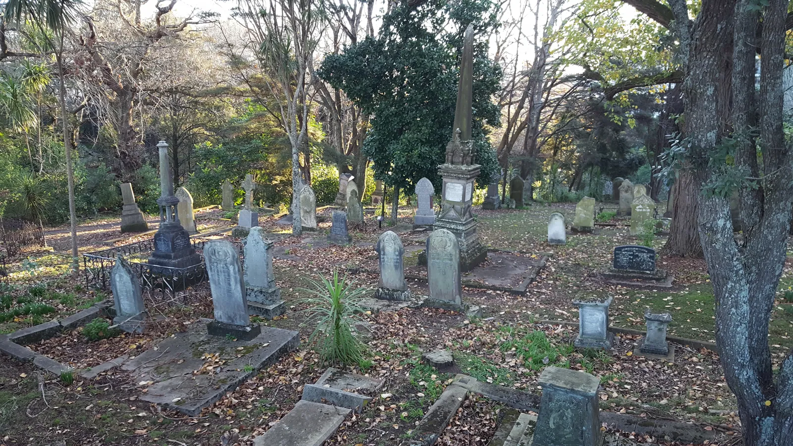 Weathered nineteenth-century headstones standing among trees and fallen leaves in Symonds Street Cemetery, Auckland